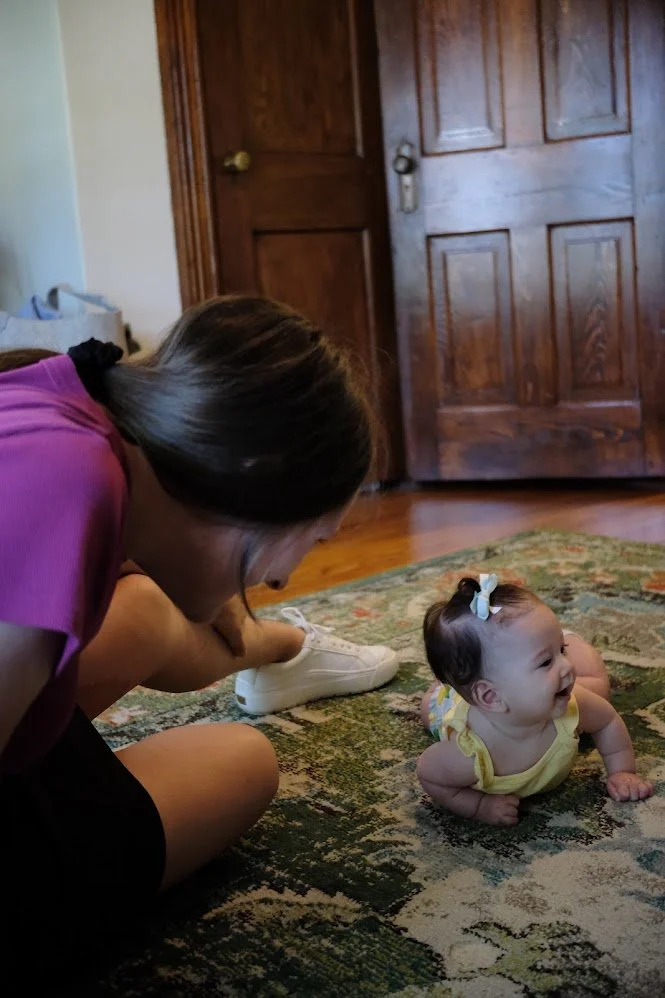 A woman with long brown hair in a pink shirt is sitting on a patterned rug on the floor, playing with a smiling baby girl wearing a yellow outfit and a white bow in her hair. They are in front of wooden doors in a room.