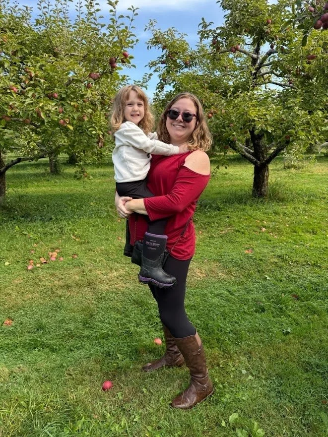 A woman holding a young girl in an apple orchard with trees full of apples. They are smiling, and the woman is wearing sunglasses, a red top with open shoulders, black leggings, and brown boots. The girl is wearing a white hoodie, black pants, and has blonde hair.