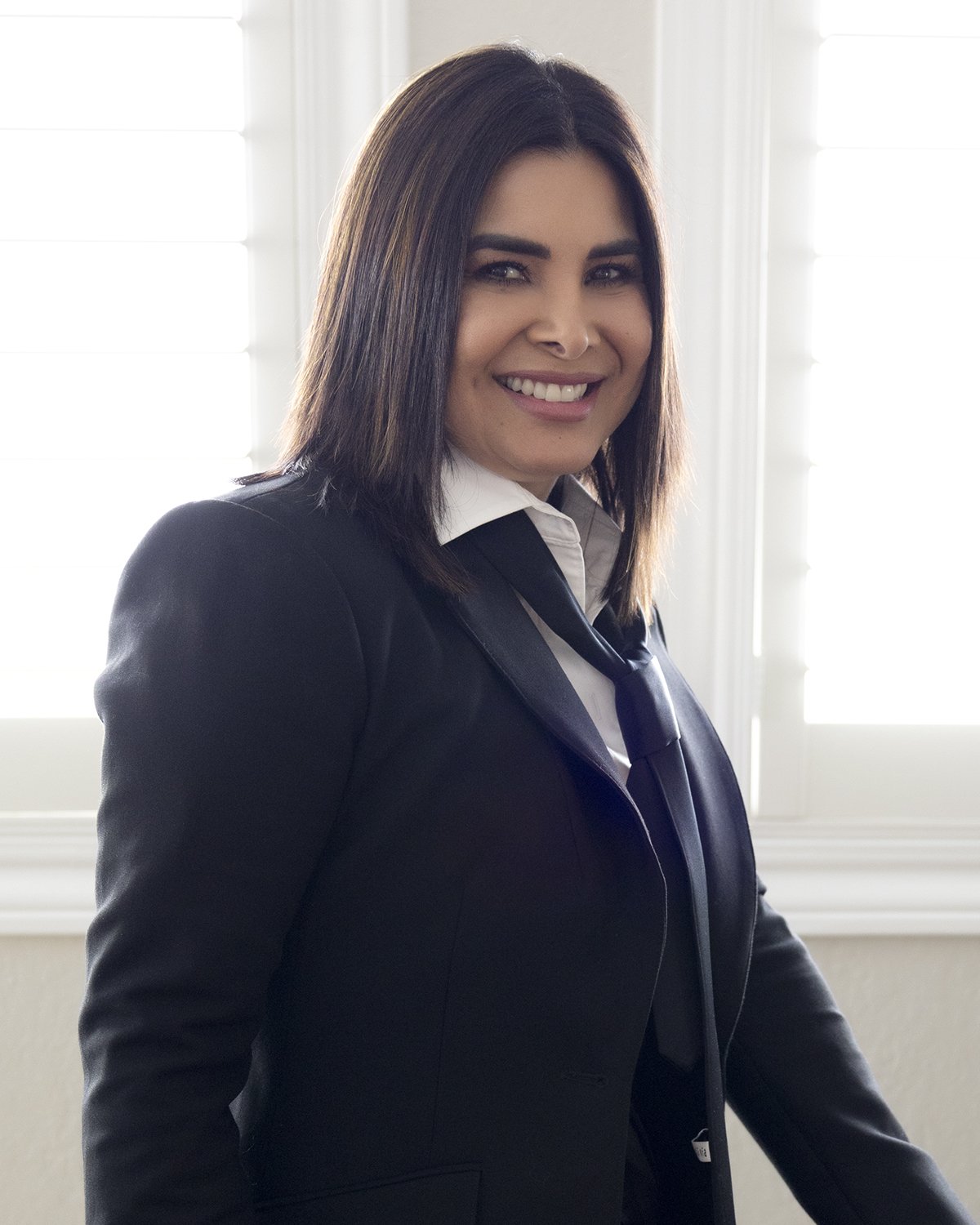 A woman with shoulder-length dark hair in a black blazer, white shirt, and black tie, standing in front of a bright window.