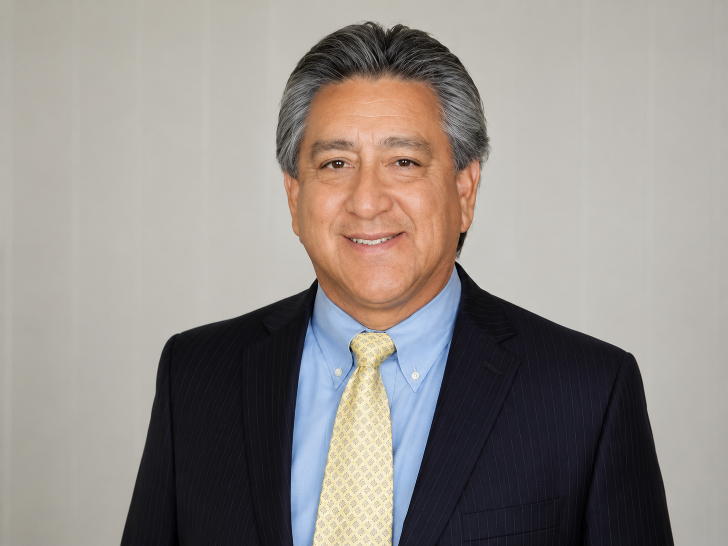 A middle-aged man with gray hair, smiling, wearing a dark pinstripe suit, light blue shirt, and yellow tie, standing against a neutral background.