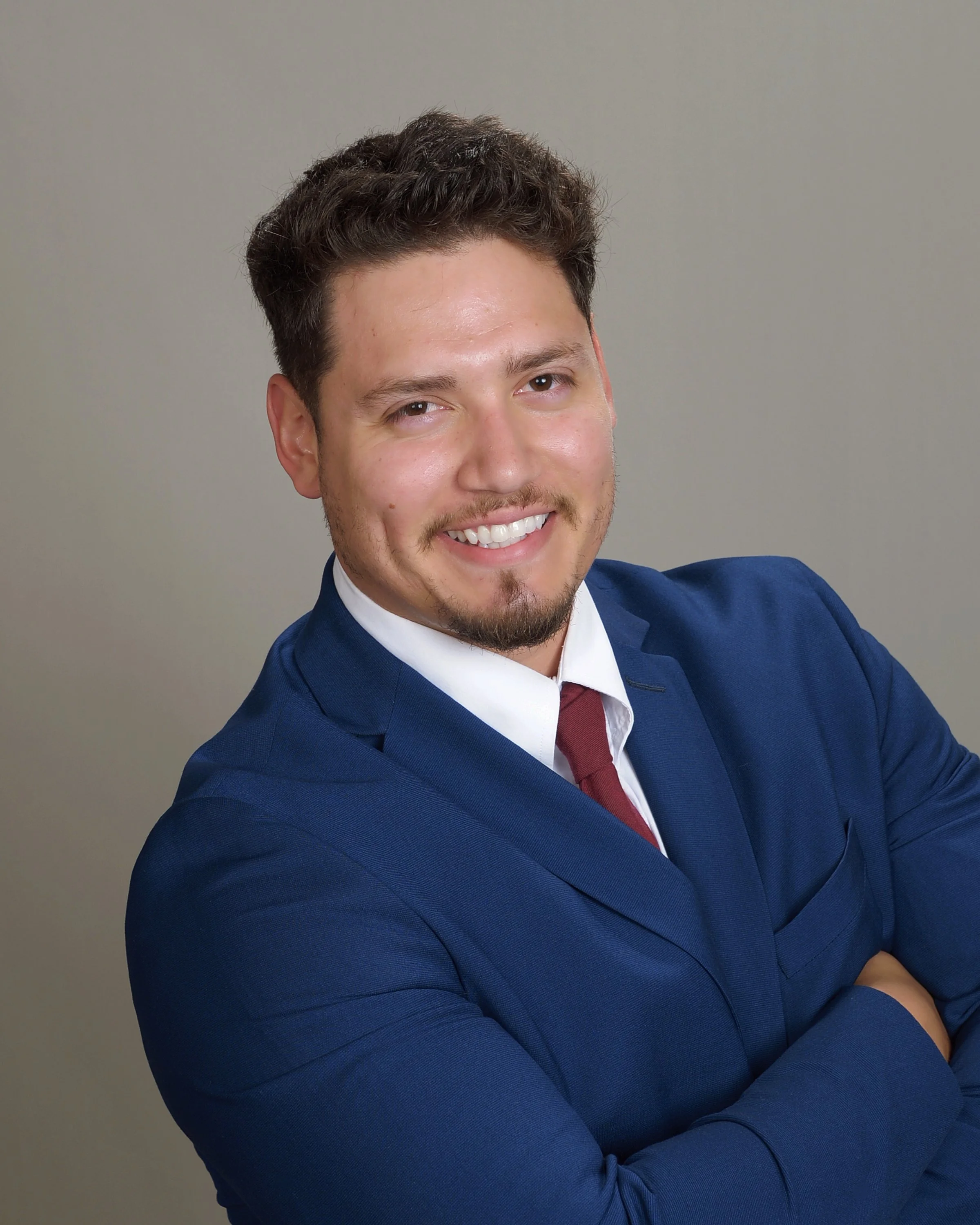 Close-up of a man smiling, wearing a navy blue suit, white shirt, and red tie, with arms crossed, in front of a plain gray background.