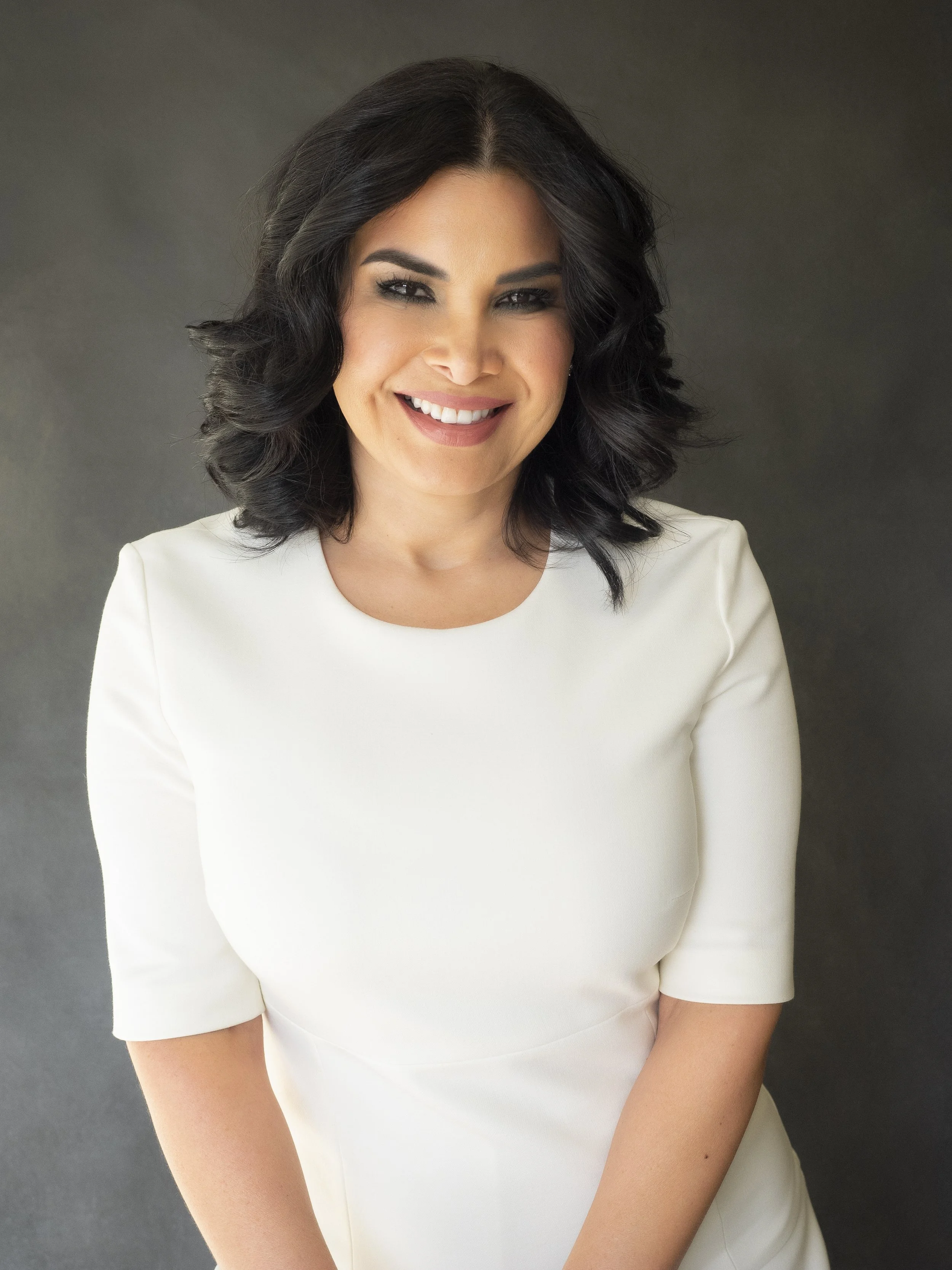 A woman with dark, curly hair and a bright smile, wearing a white dress against a dark background.