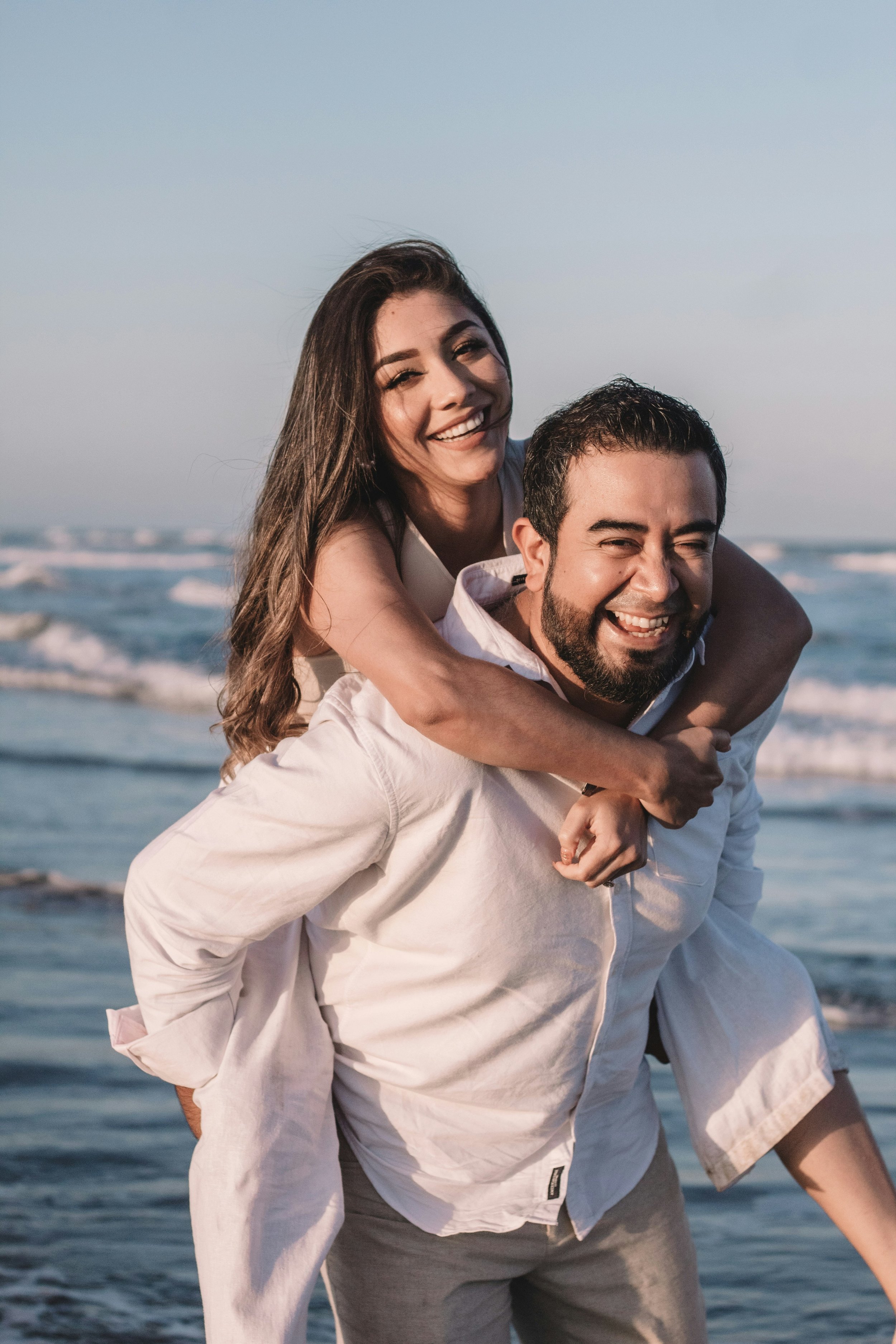A smiling woman with long brown hair riding on the back of a man with short dark hair and a beard at the beach, both wearing light-colored clothing, with the ocean and sky in the background.