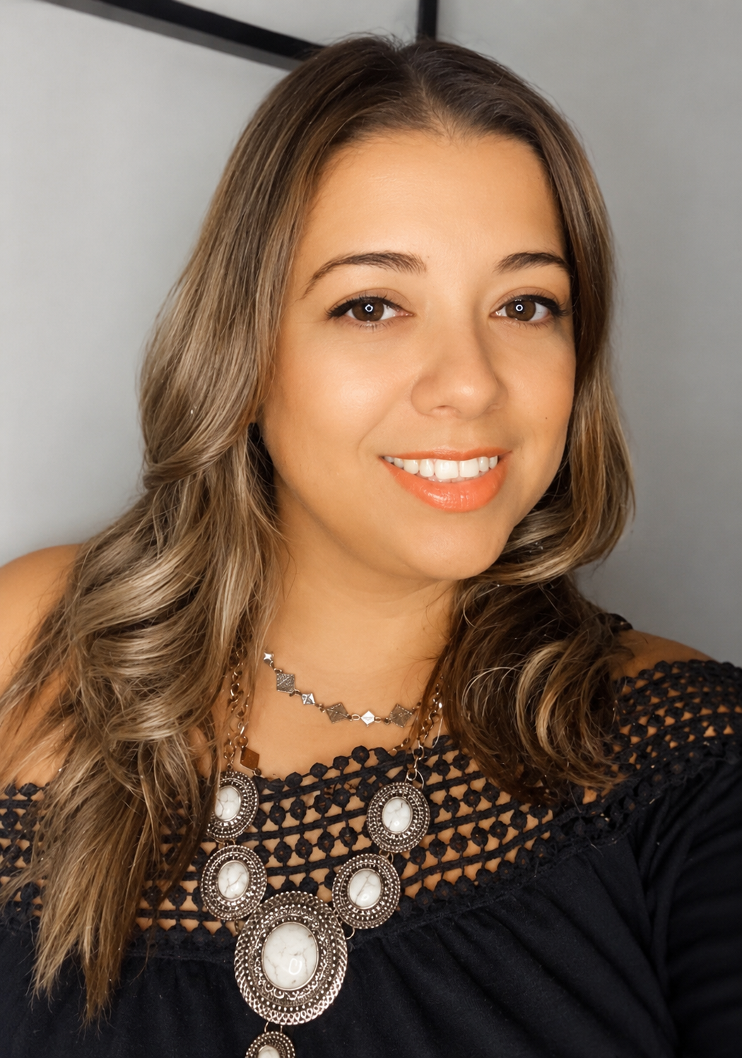 A woman with long, wavy brown hair smiling at the camera, wearing an intricate black top with crochet details and a large statement necklace with white stones.