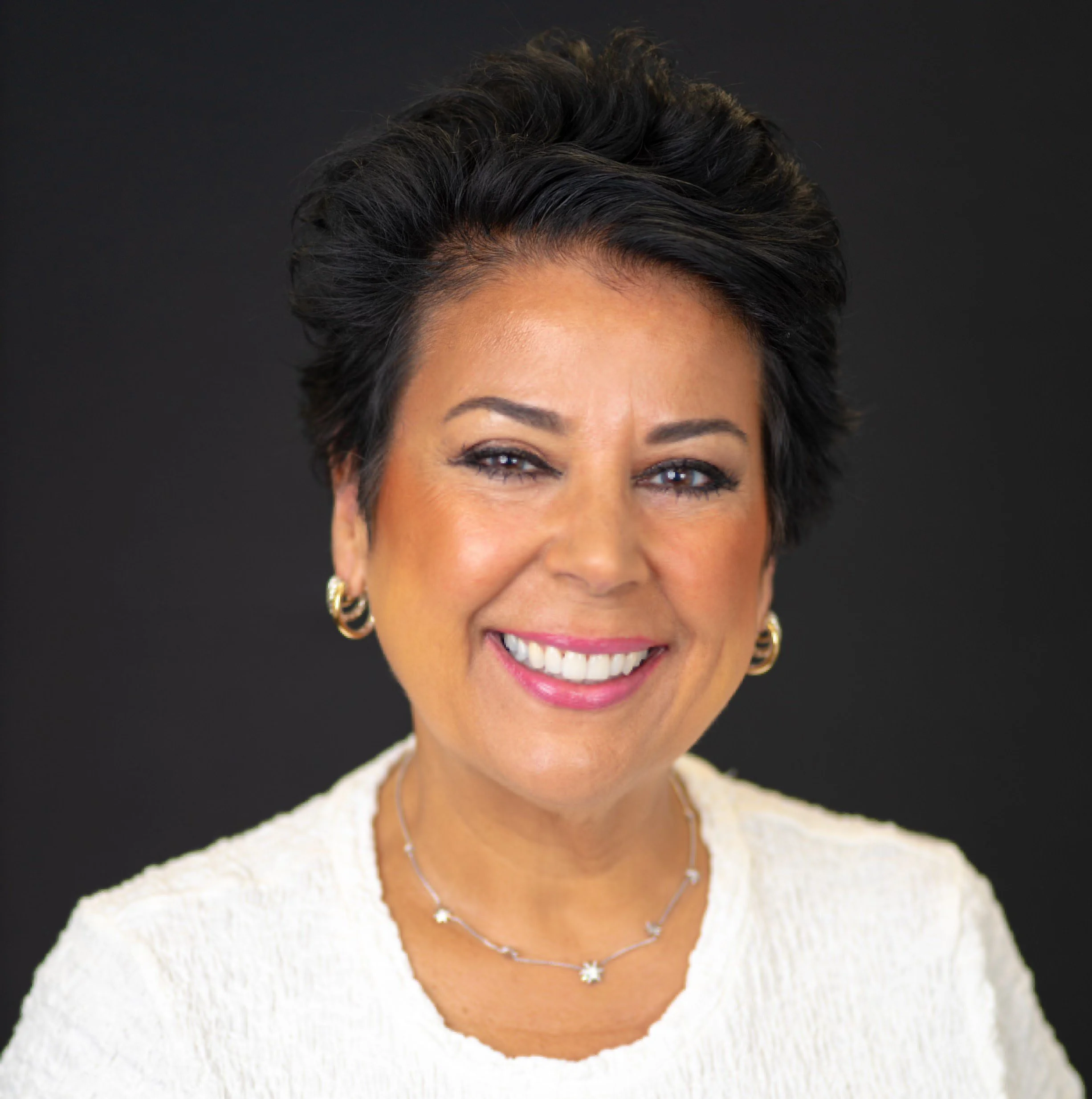 Close-up of a smiling woman with short dark hair, wearing gold earrings, a silver necklace, and a white textured top, set against a black background.