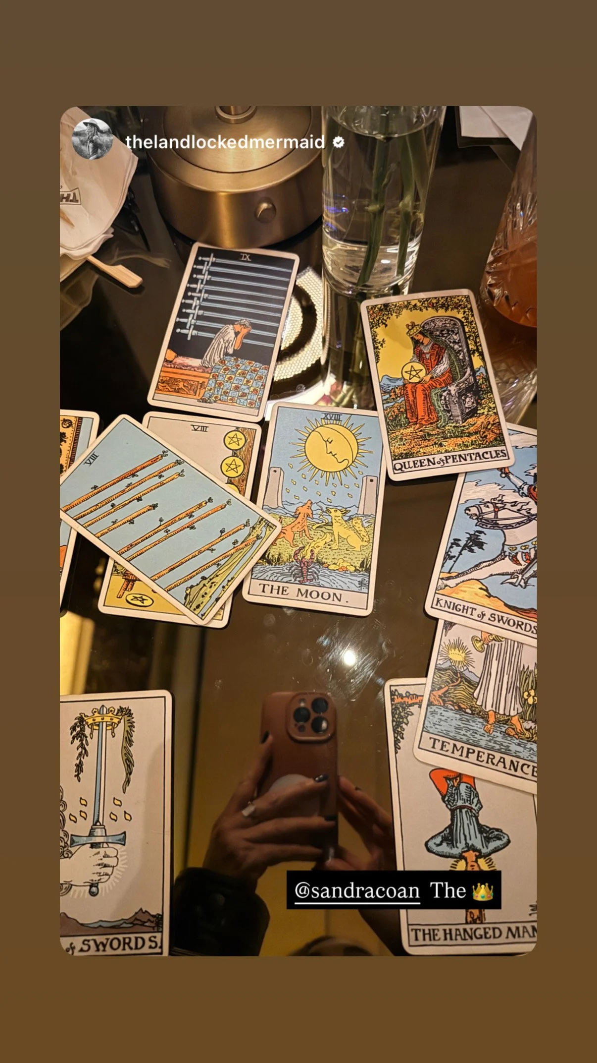 Tarot cards spread out on a table, including The Moon, Queen of Pentacles, Knight of Swords, and Temperance. There are also water glasses, a flower vase, and a gold-colored object on the table.