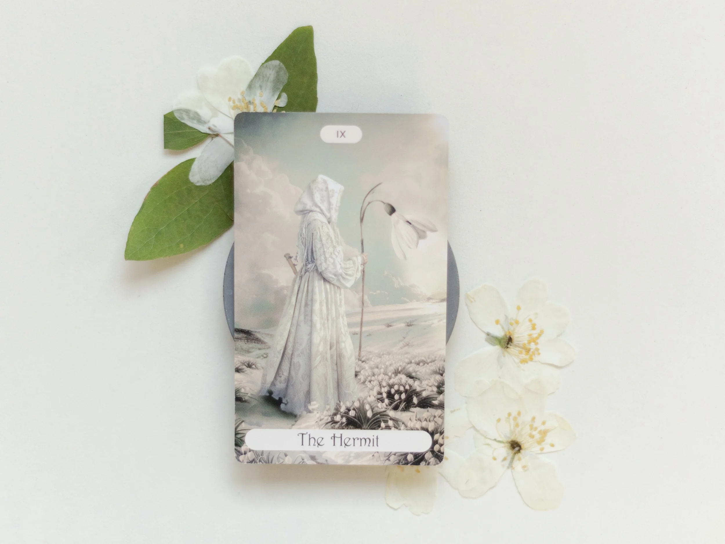 A tarot card titled The Hermit featuring a woman in a white dress holding a staff, with white flowers and green leaves surrounding it, and some white blossoms with yellow stamens nearby.