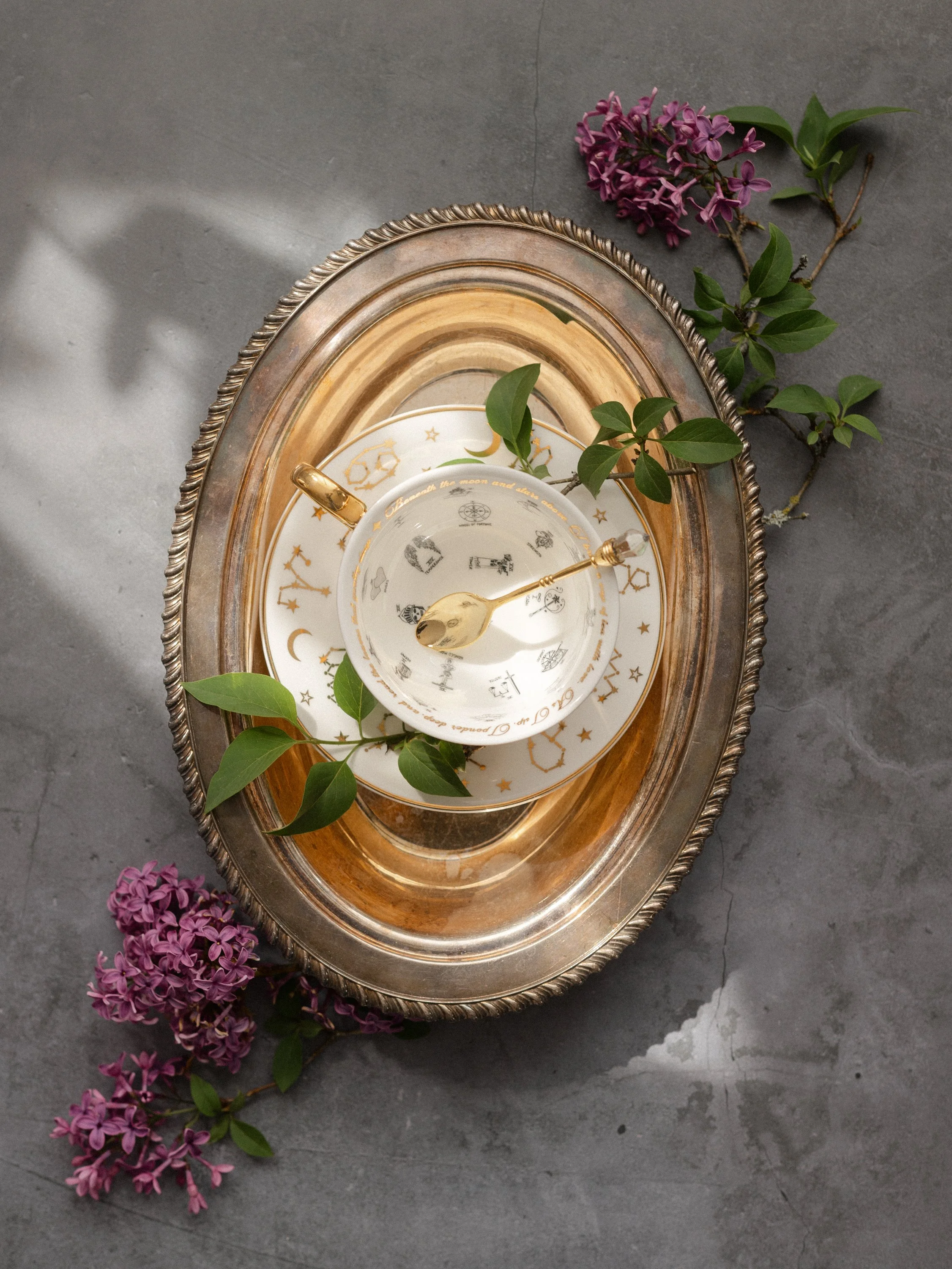Decorative oval silver tray with gold-accented celestial-themed tea set, surrounded by purple lilac flowers and green leaves on a gray surface.