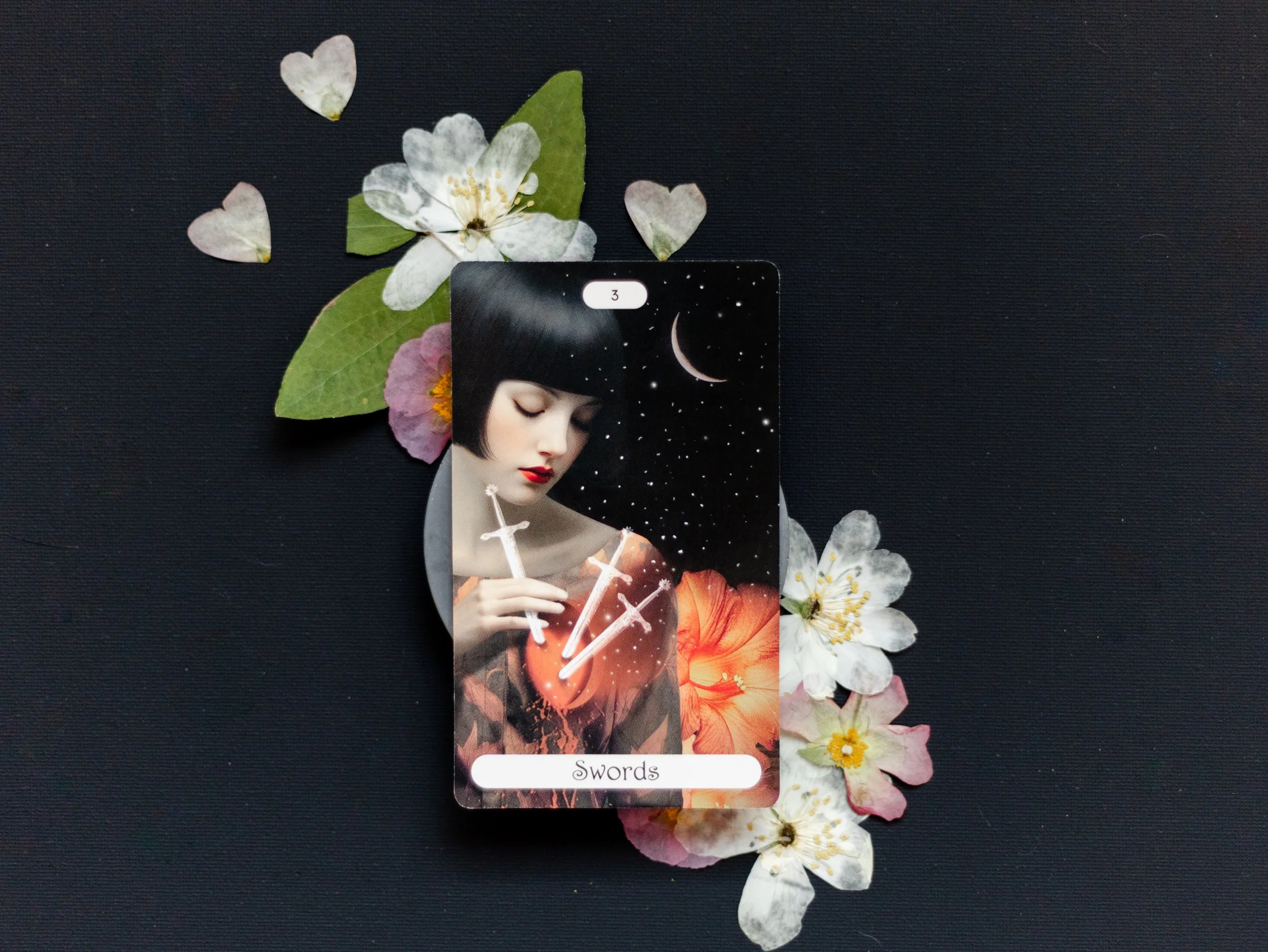 A tarot card labeled 'Swords' is placed on a black surface, surrounded by white and pink flower petals. The card depicts a woman with short dark hair and a serious expression, holding two glowing swords against a starry night sky with a crescent moon.
