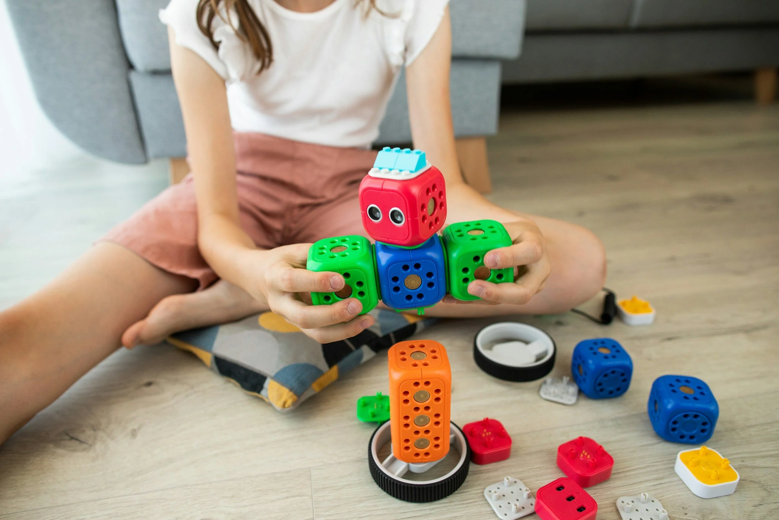 A young girl sits cross-legged on the floor playing with colorful building blocks and toy dice, building a miniature structure. She is wearing a white shirt and pink shorts, with a gray couch visible in the background.