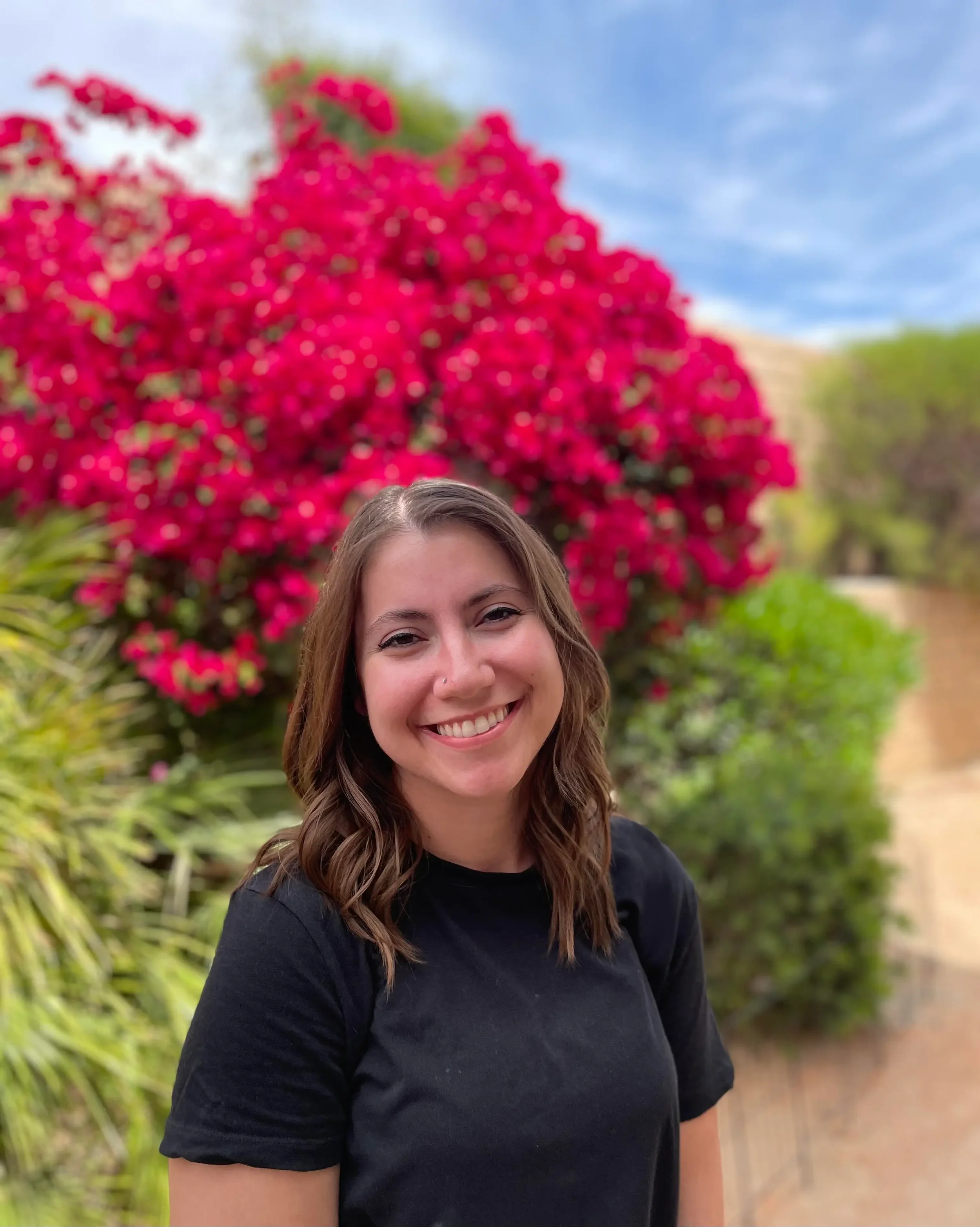 A young woman smiling outdoors, with shoulder-length wavy hair, wearing a black shirt, standing in front of a vibrant pink flowering bush and green foliage under a partly cloudy sky.