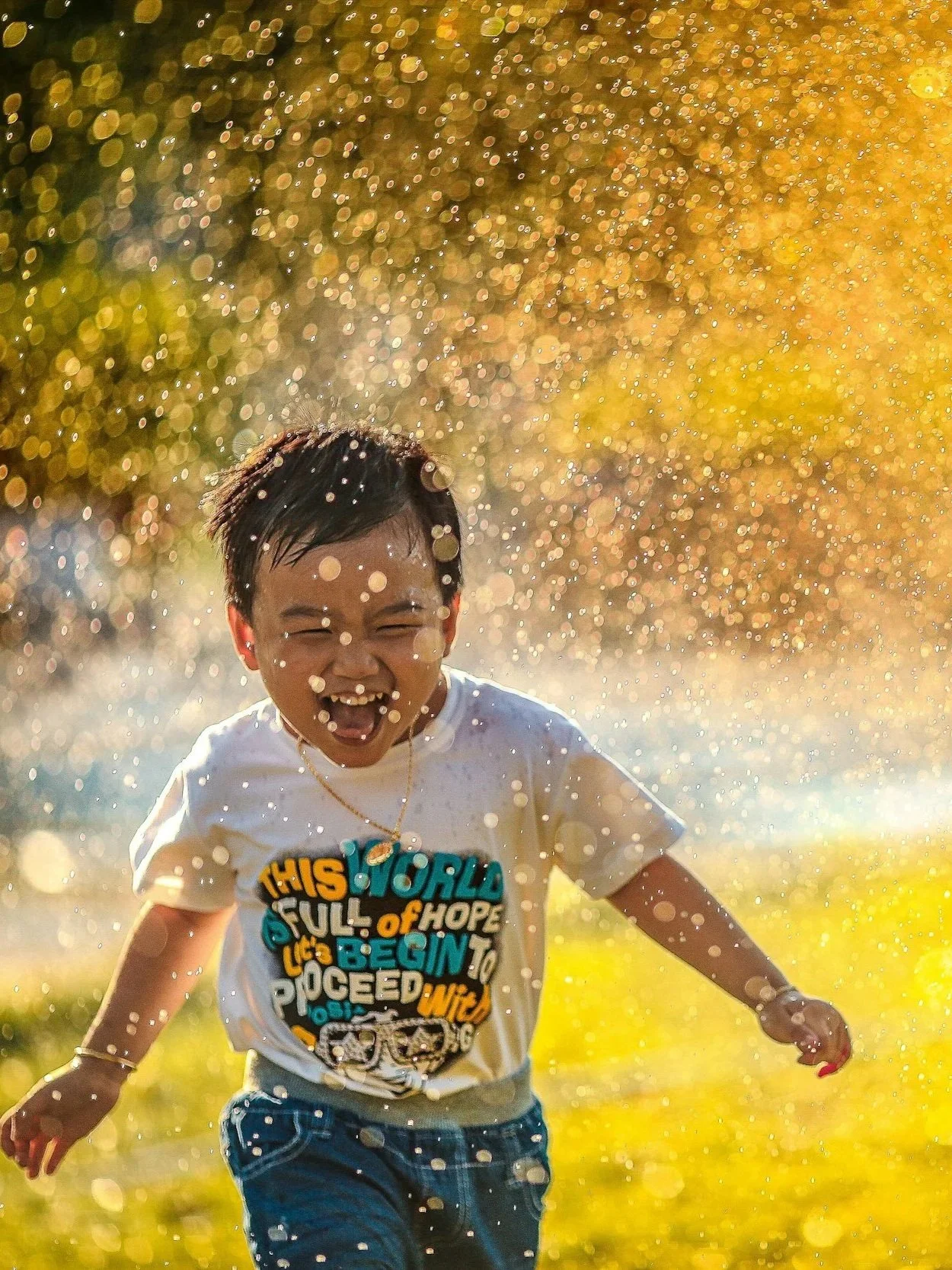 A child playing and laughing while running through a water spray, with sunlight creating a bokeh effect in the background.