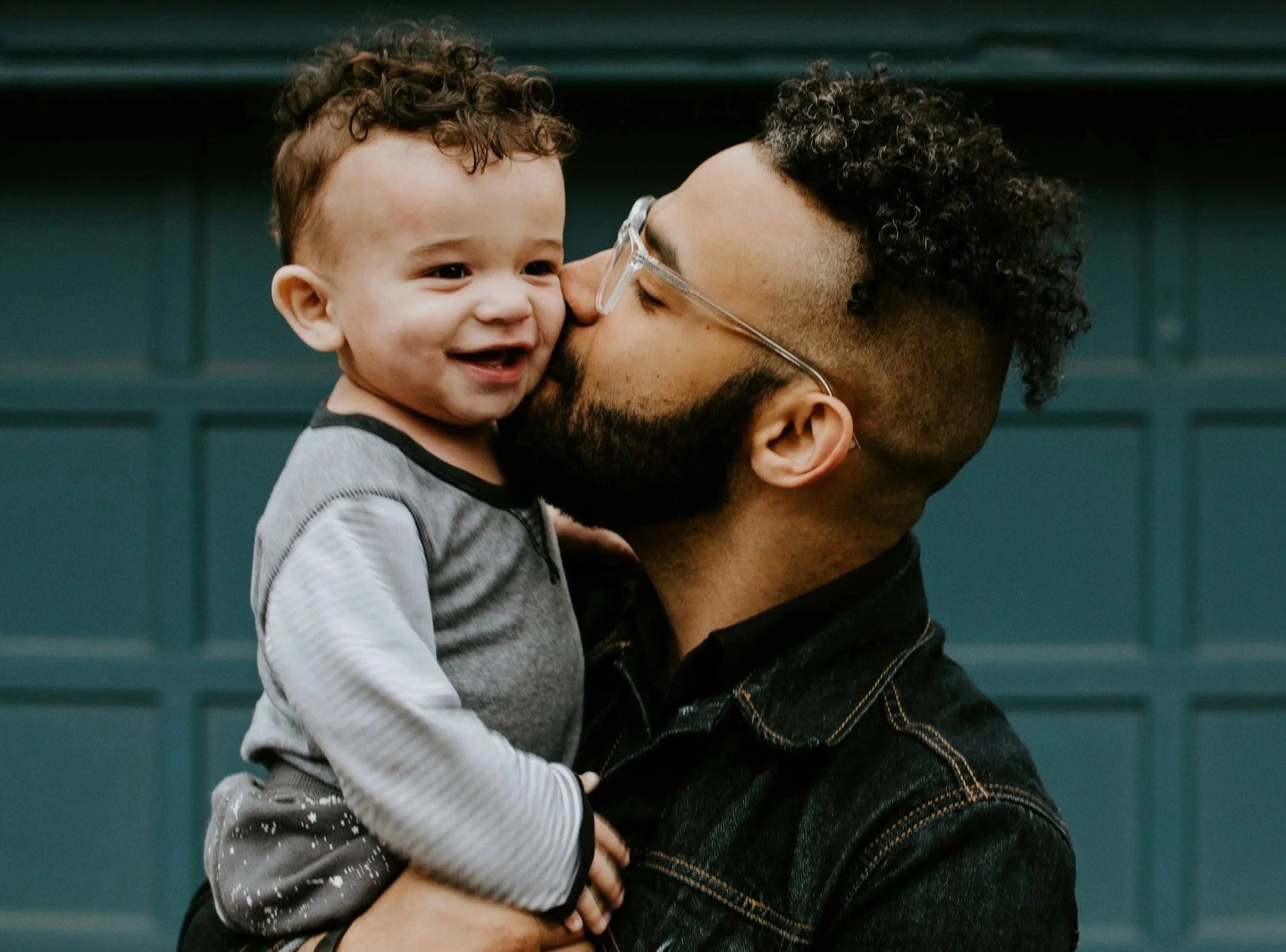 A man with glasses and a beard holding a young child in his arms, both sharing a joyful and loving moment with their faces close together, against a teal-colored background.