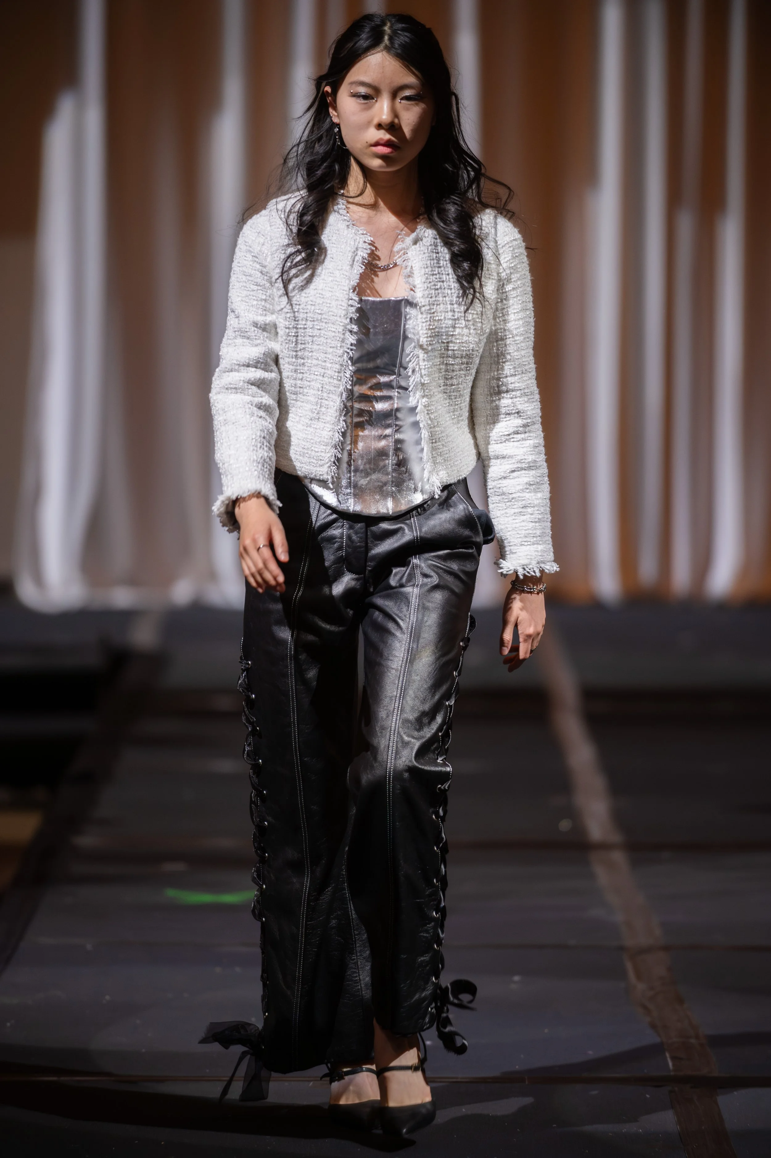 A woman walking on a runway during a fashion show, wearing a white textured jacket, a shiny silver top, and black leather pants with ribbon details.