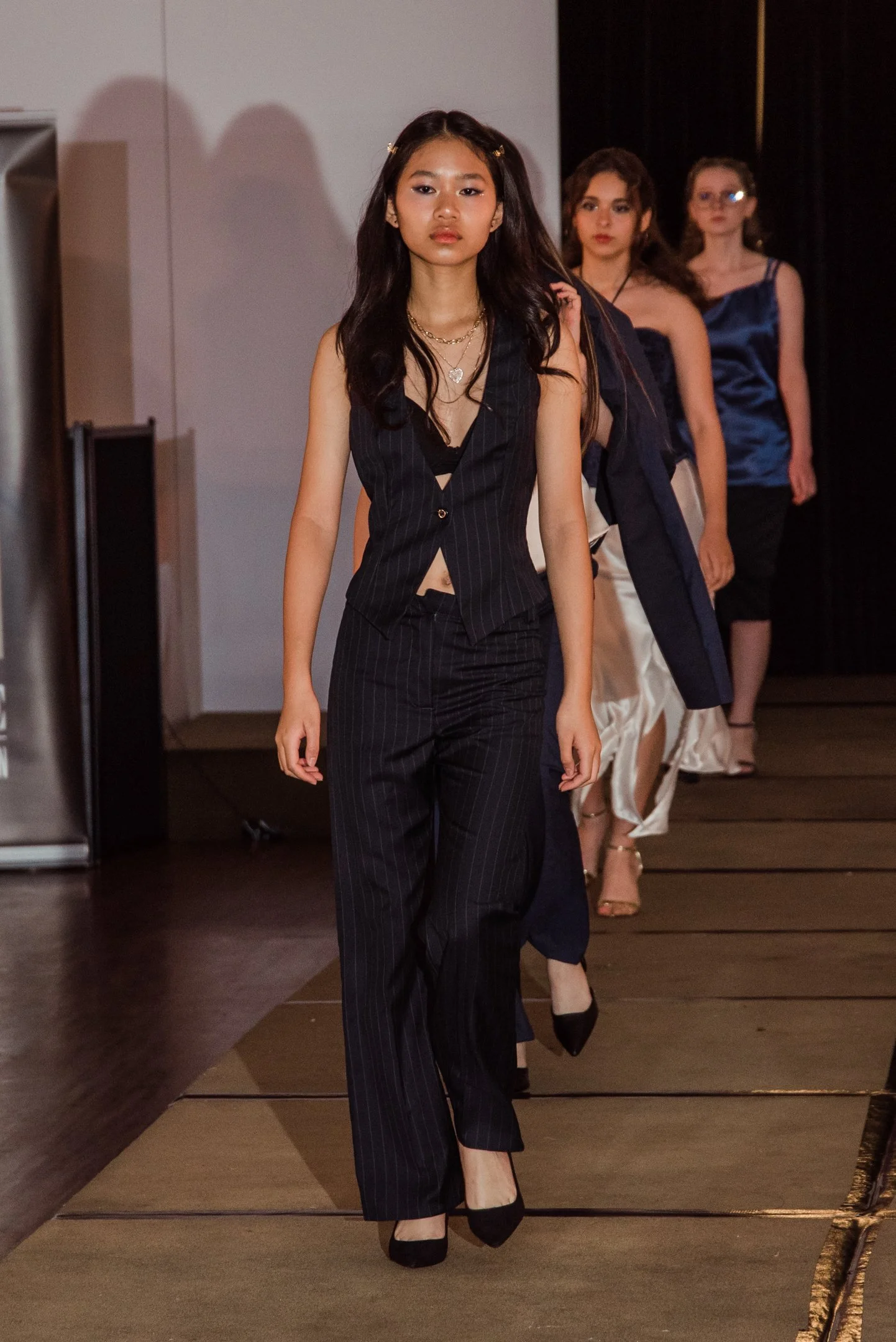 A fashion runway with four models walking in a line. The model in the front wears a black pinstripe suit with black heels and layered necklaces. The other models follow wearing elegant outfits, including satin dresses and suits, with a dark background and a carpeted runway.