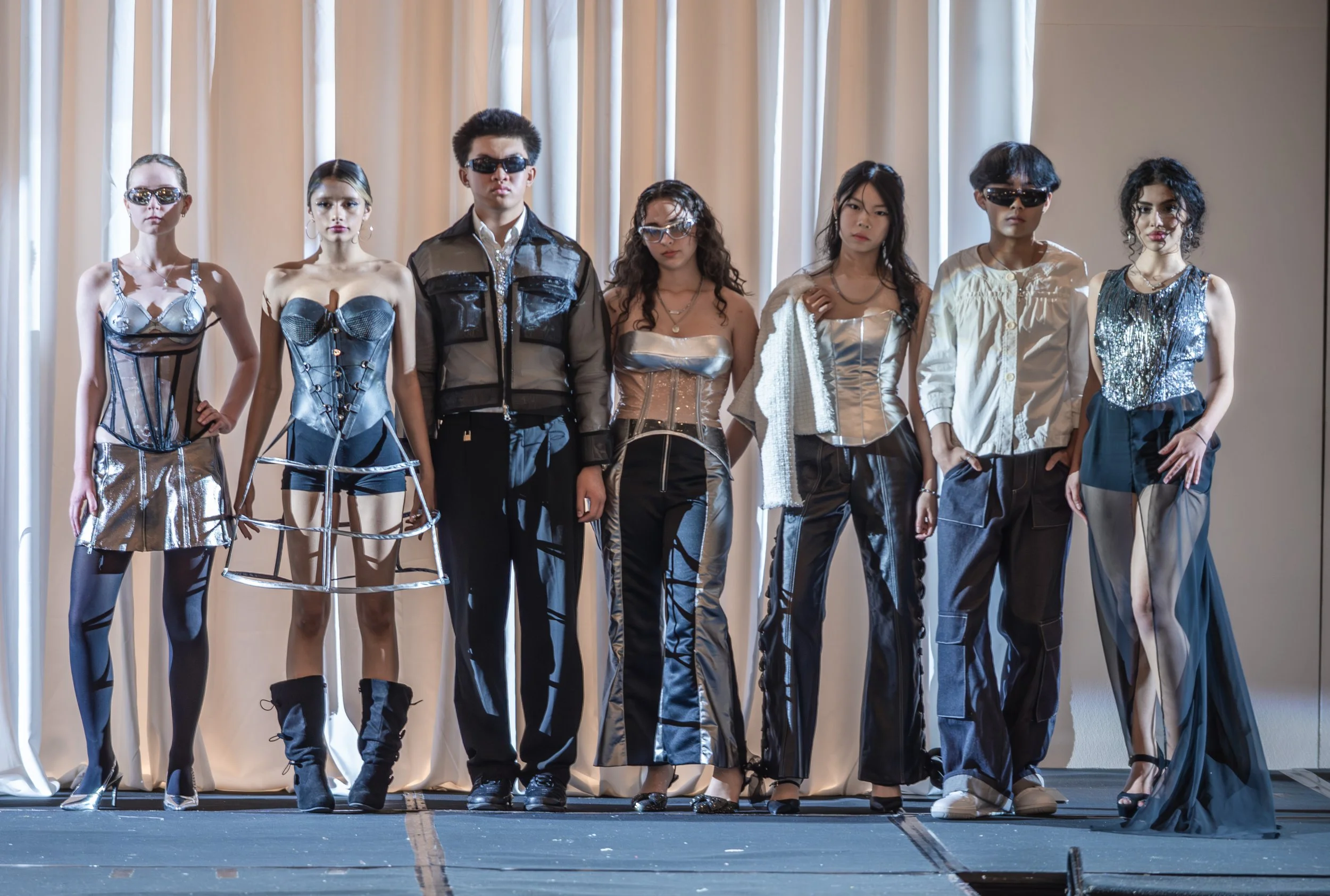 Group of seven young models standing on a runway stage, wearing futuristic and edgy fashion clothing with metallic, plastic, and leather materials, in front of a beige curtain.