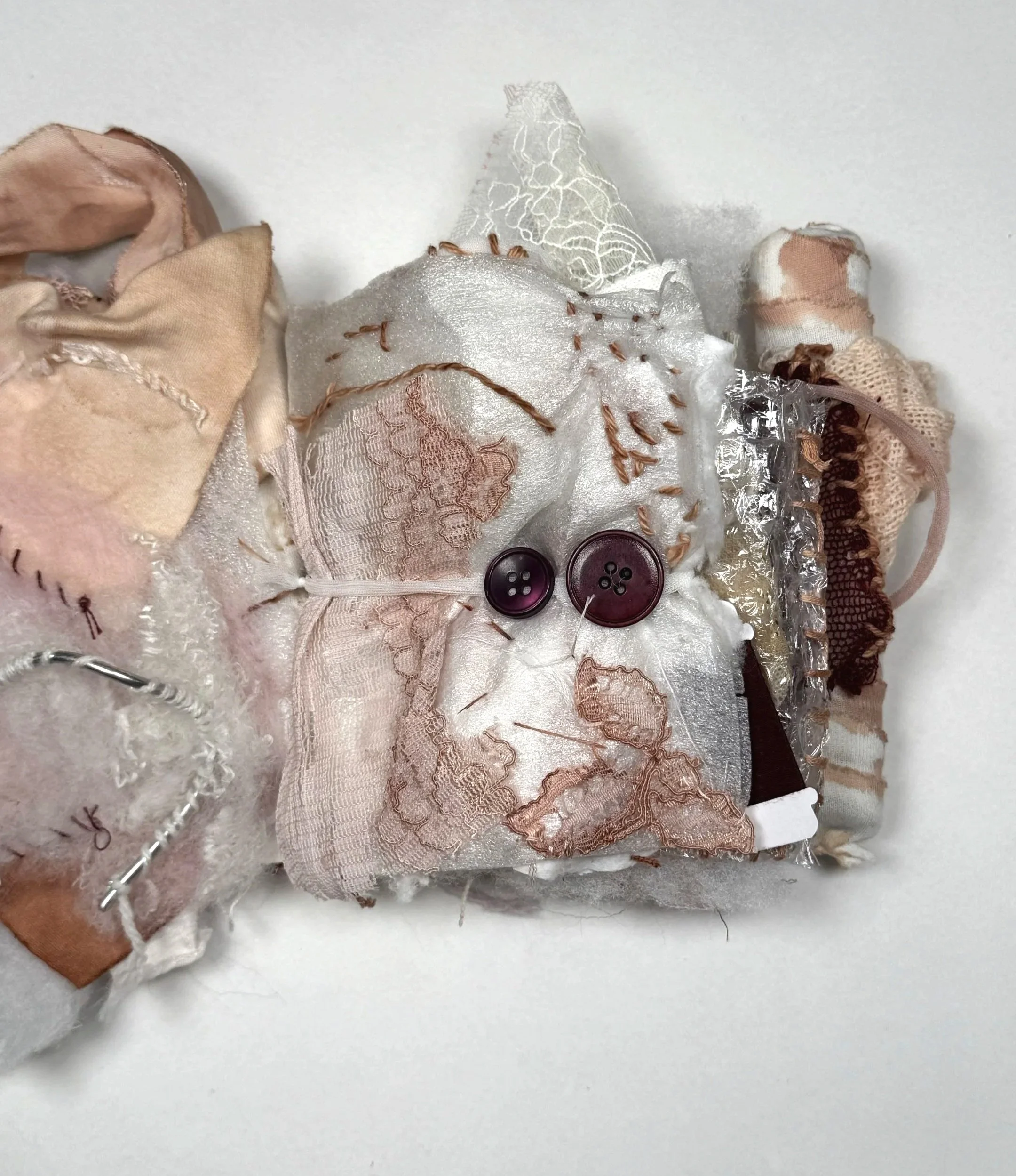 Close-up of a doll made from fabric and lace, featuring stitched details, ribbons, buttons, and ribbons on a white background.