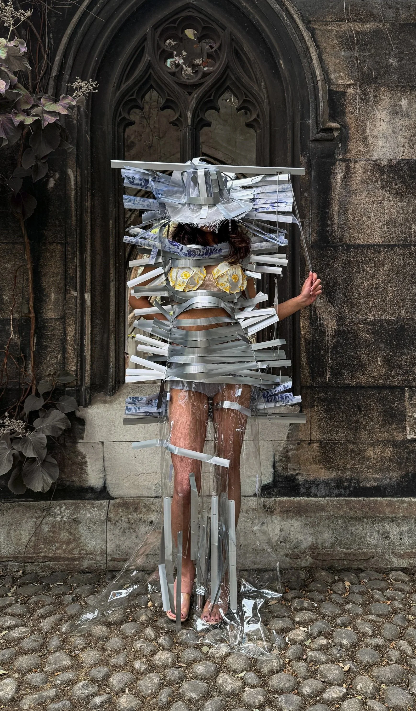 A person is standing against a stone wall in front of a gothic-style arched window. Model wearing garment and hat made out of aluminium window blinds, with yellow floral bra top peaking from under.