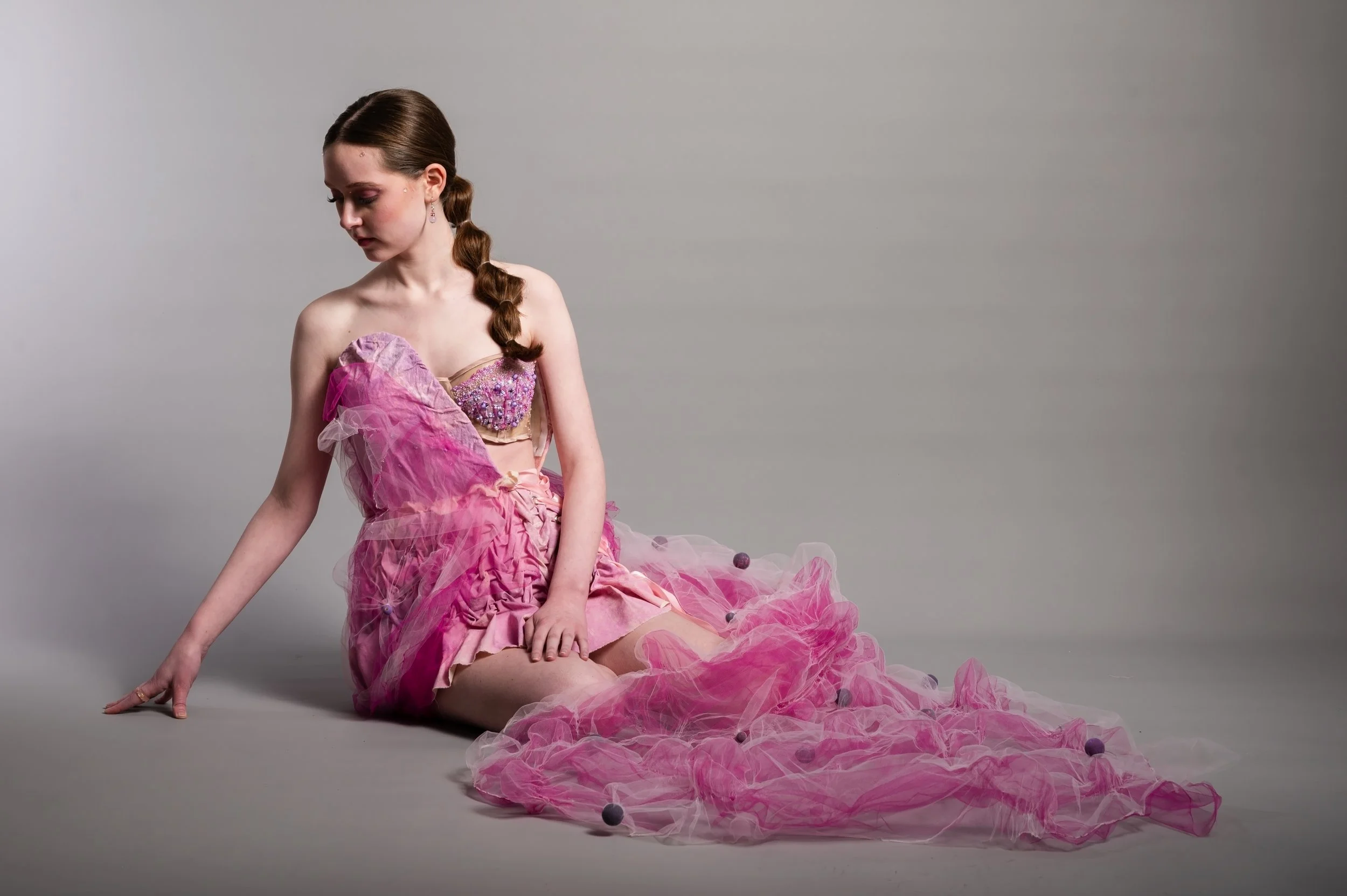 A young woman with long brown hair styled in a braid, wearing a pink dress with intricate beading on the bodice and layers of pink tulle, is sitting on the floor against a plain gray background, gazing downward with a contemplative expression.