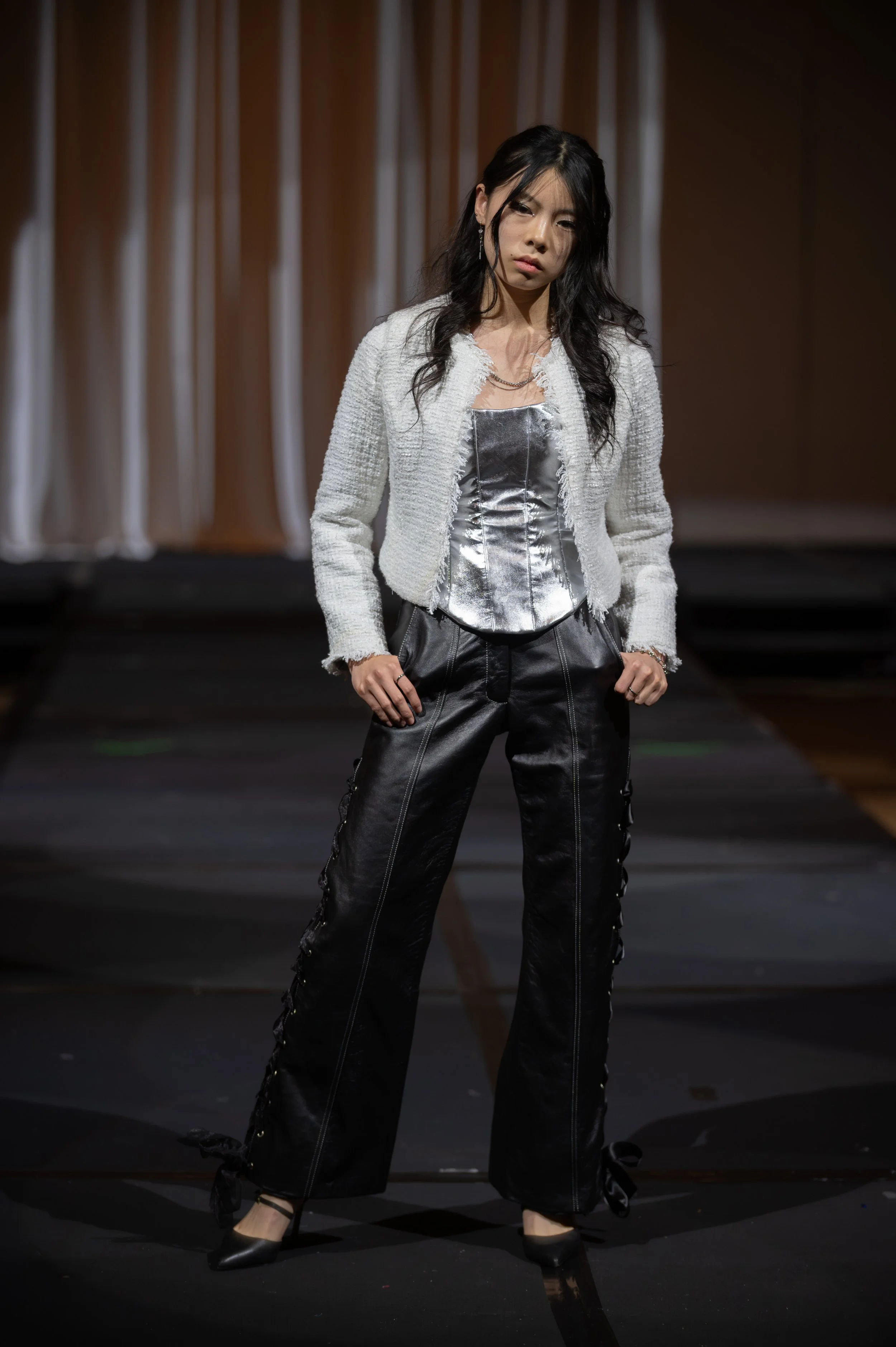 A woman with long black hair and light skin standing on a stage with a dark background. She is wearing a shiny silver top, a white textured jacket, black leather pants with lace-up details on the sides, and black high-heeled shoes.