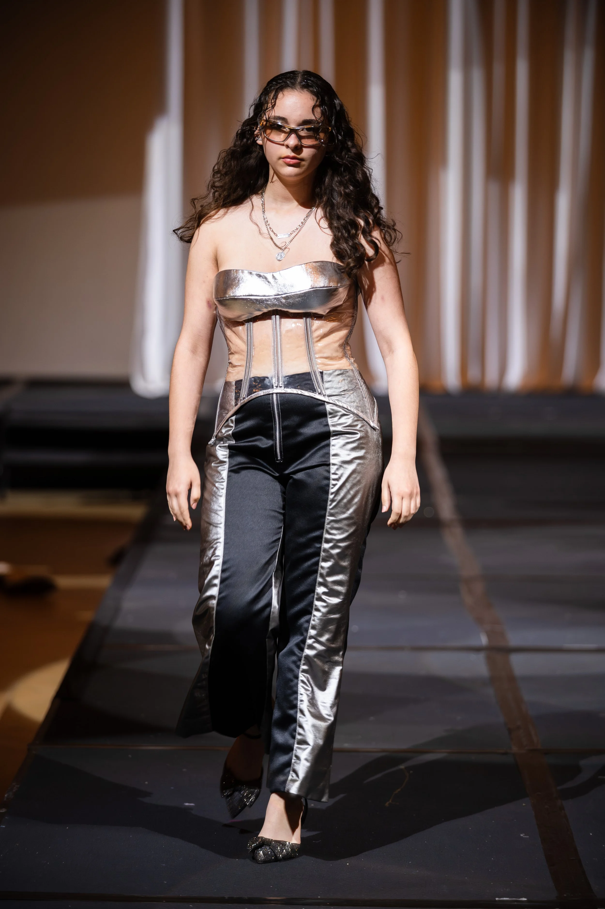 A woman walking on a runway during a fashion show, wearing a metallic strapless top and matching pants with black accents, black shoes with glitter details, glasses, and layered necklaces.
