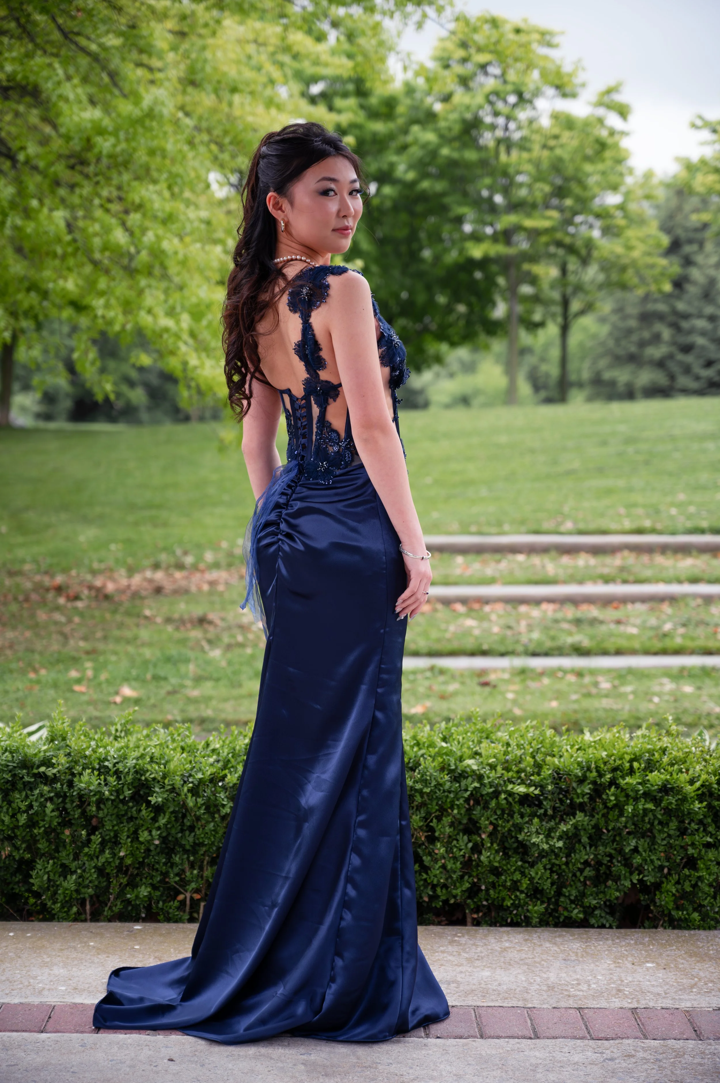A woman in an elegant navy blue evening gown standing outdoors in a park with green trees and bushes in the background.