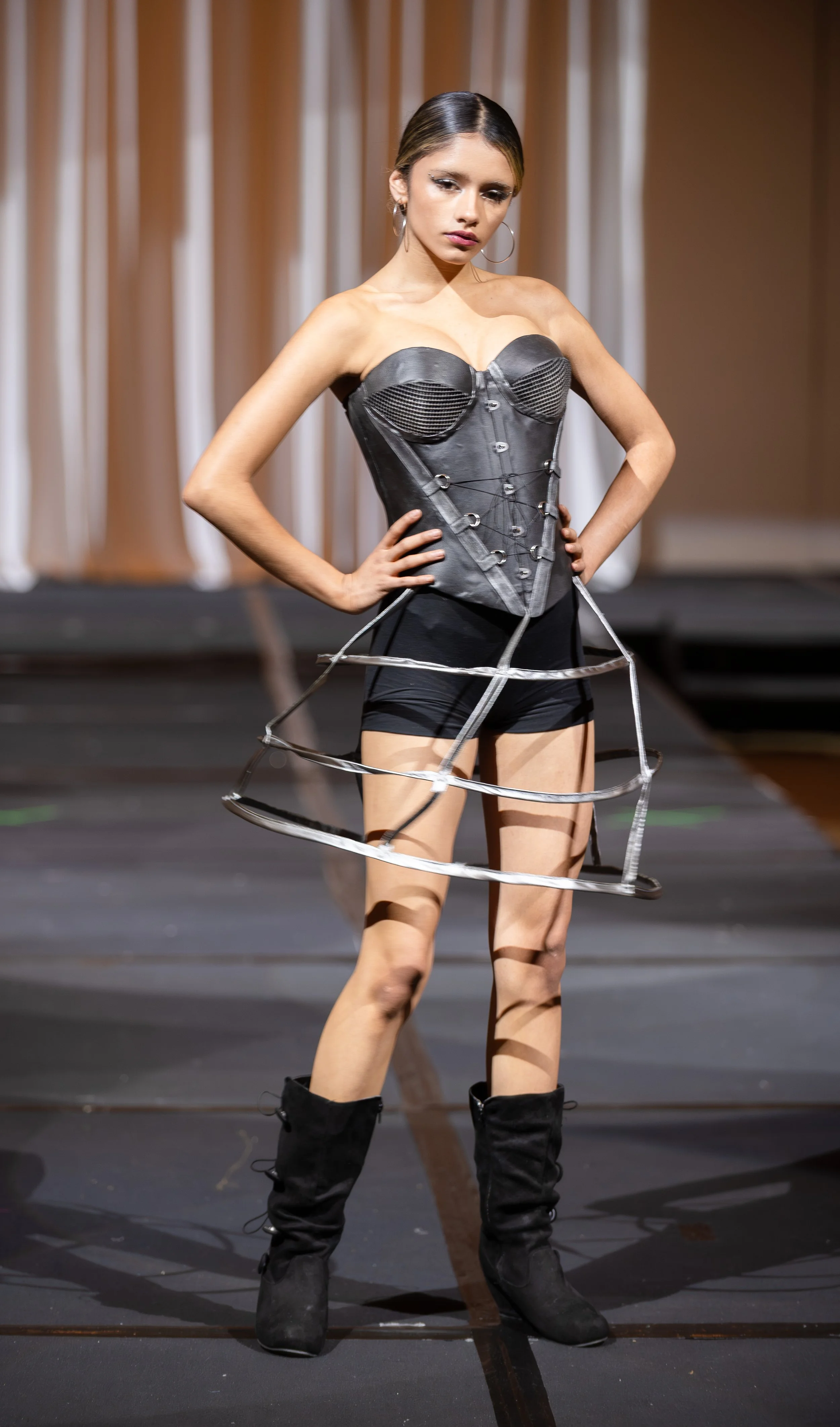 A woman on a runway dressed in a cast-iron corset with metal hoops around her waist and black boots, in front of curtains.