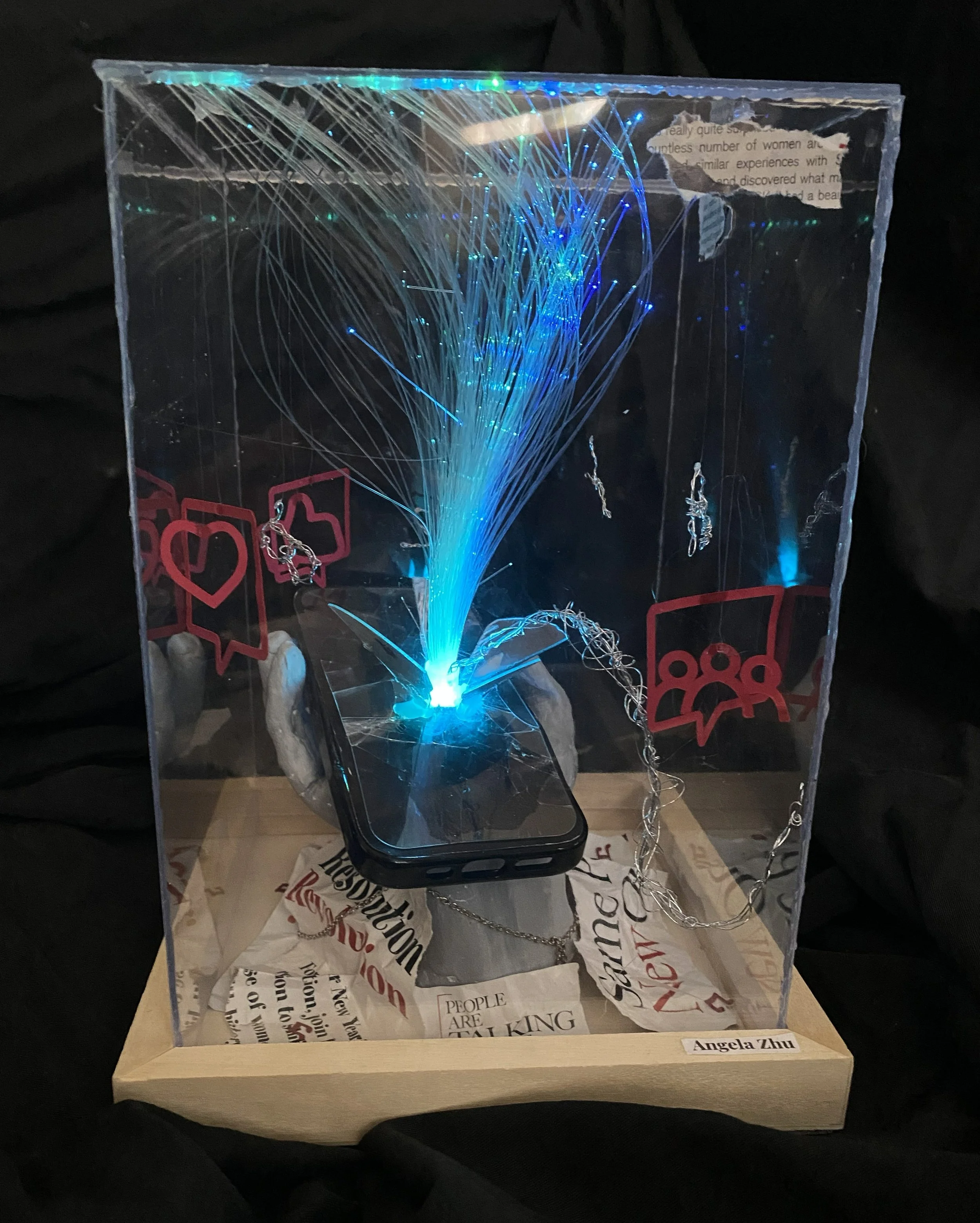 A display case with blue fiber optic branches illuminated inside, surrounded by paper with text and jewelry pieces, on a wooden stand with a name tag reading 'Angela Zhu'.