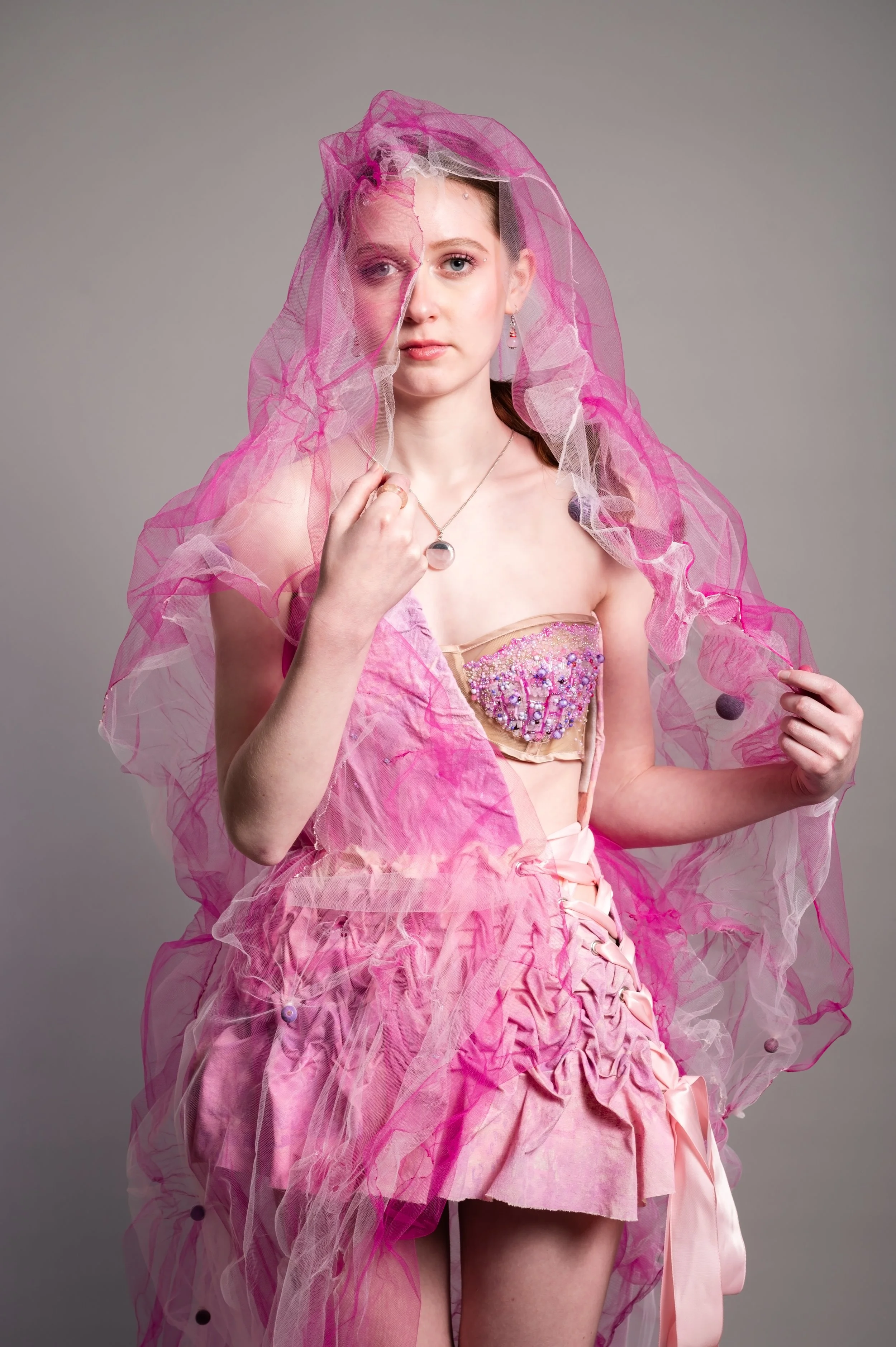 A woman wearing a pink dress and a sheer pink veil with purple balls, holding the veil with one hand and wearing jewelry, against a plain gray background.