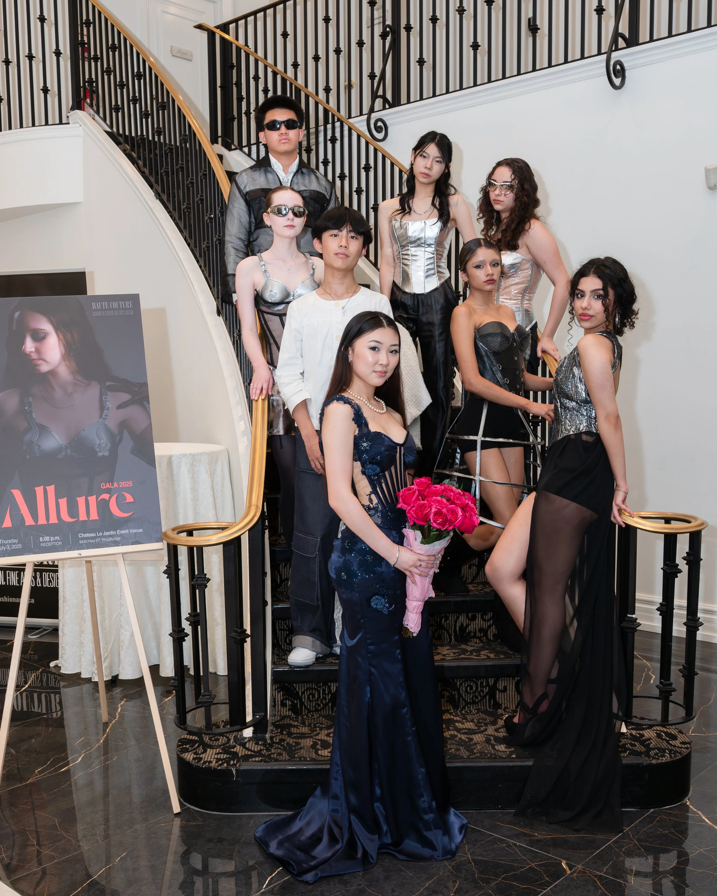 A group of diverse young people, dressed in fashionable attire, posing on a staircase at a fashion event. Some are wearing sunglasses, and one woman is holding a bouquet of pink roses. There's a poster on an easel that reads 'Allure Gala 2025'.