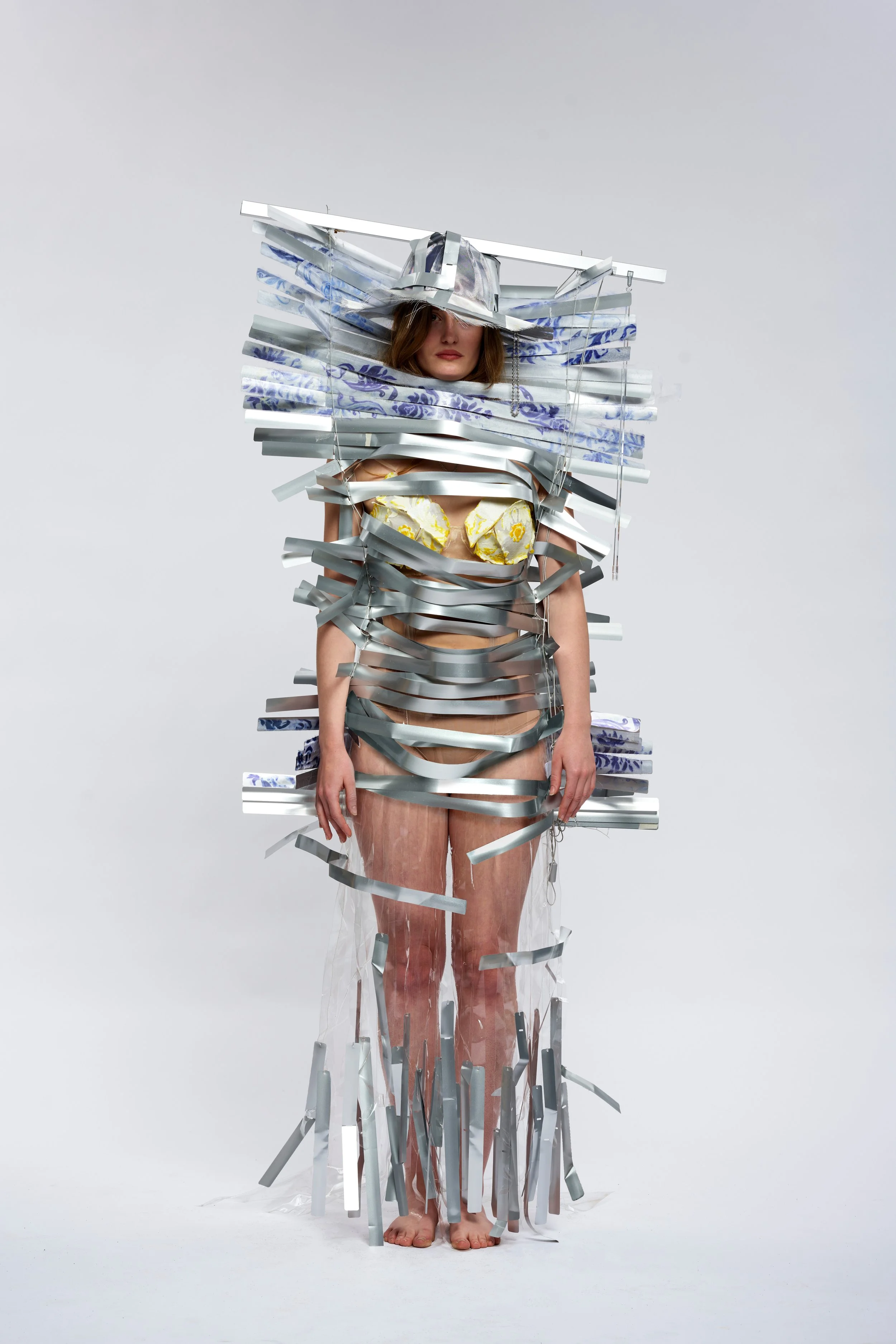 A woman standing against a plain white background, wearing garment and hat made out of aluminium window blinds, with yellow floral bra top peaking from under.