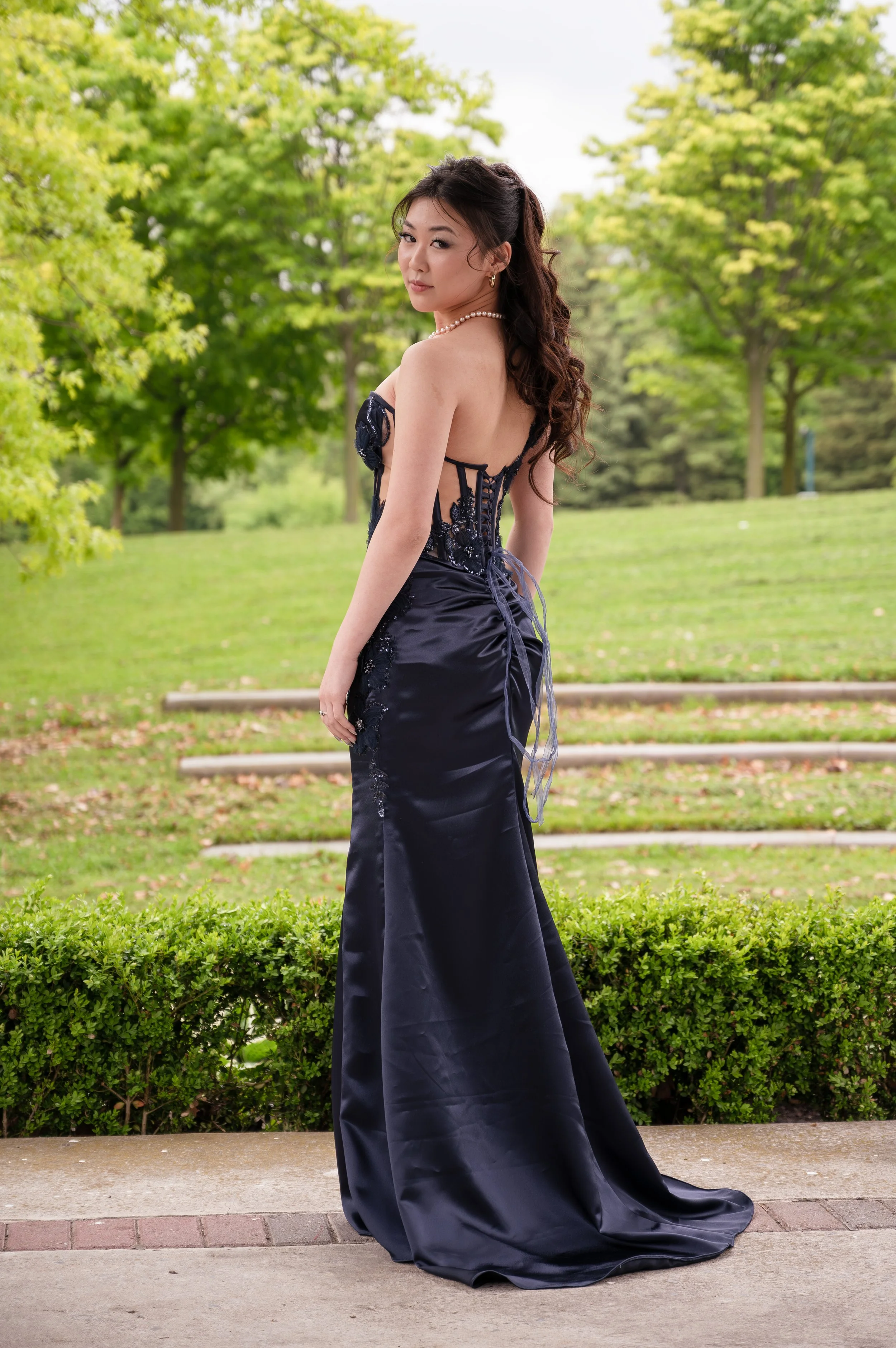 A woman in a dark blue, elegant, strapless dress with lace details, standing outdoors on a sidewalk with greenery and trees in the background. She has long, wavy hair, and is wearing a pearl necklace and earrings. She is looking over her shoulder, giving a slight smile.