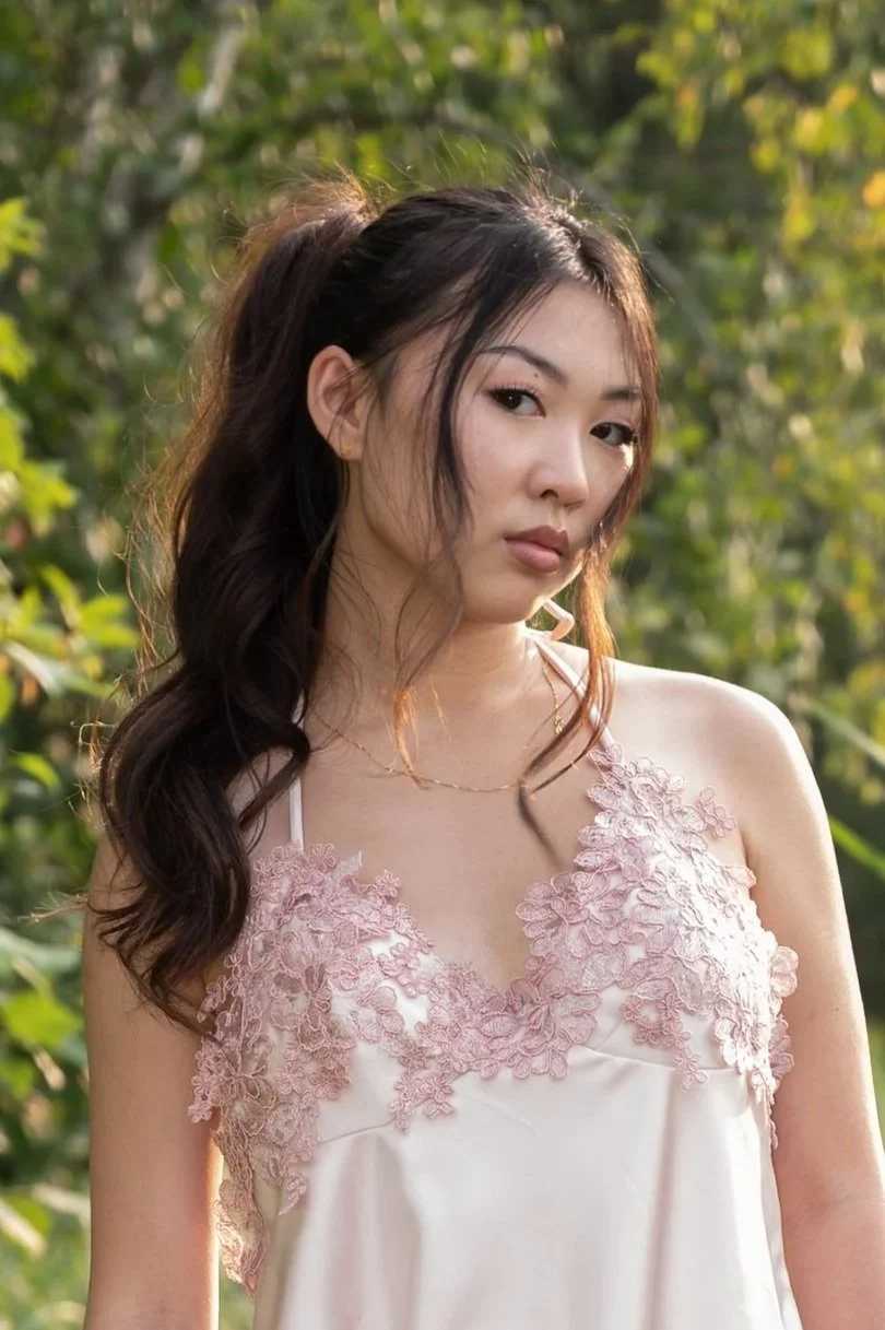 A young woman with dark hair styled in loose waves and a high ponytail stands outdoors in natural light. She wears a light pink dress with lace floral embroidery at the top. The background features green foliage.
