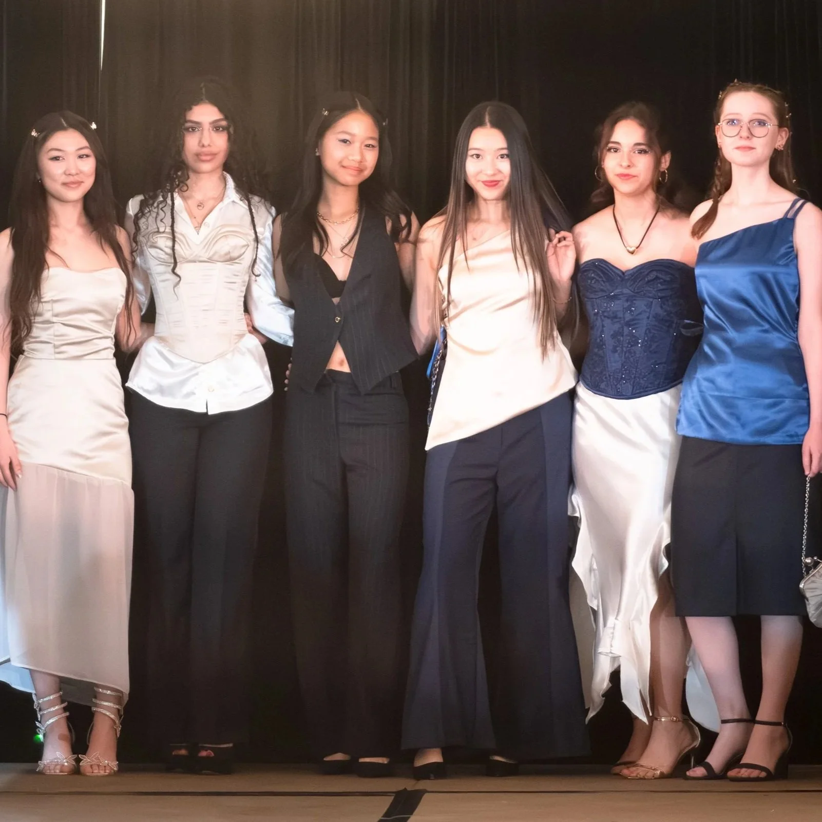 Group of seven women standing side by side on a stage, dressed in formal or semi-formal attire, with a dark curtain behind them.