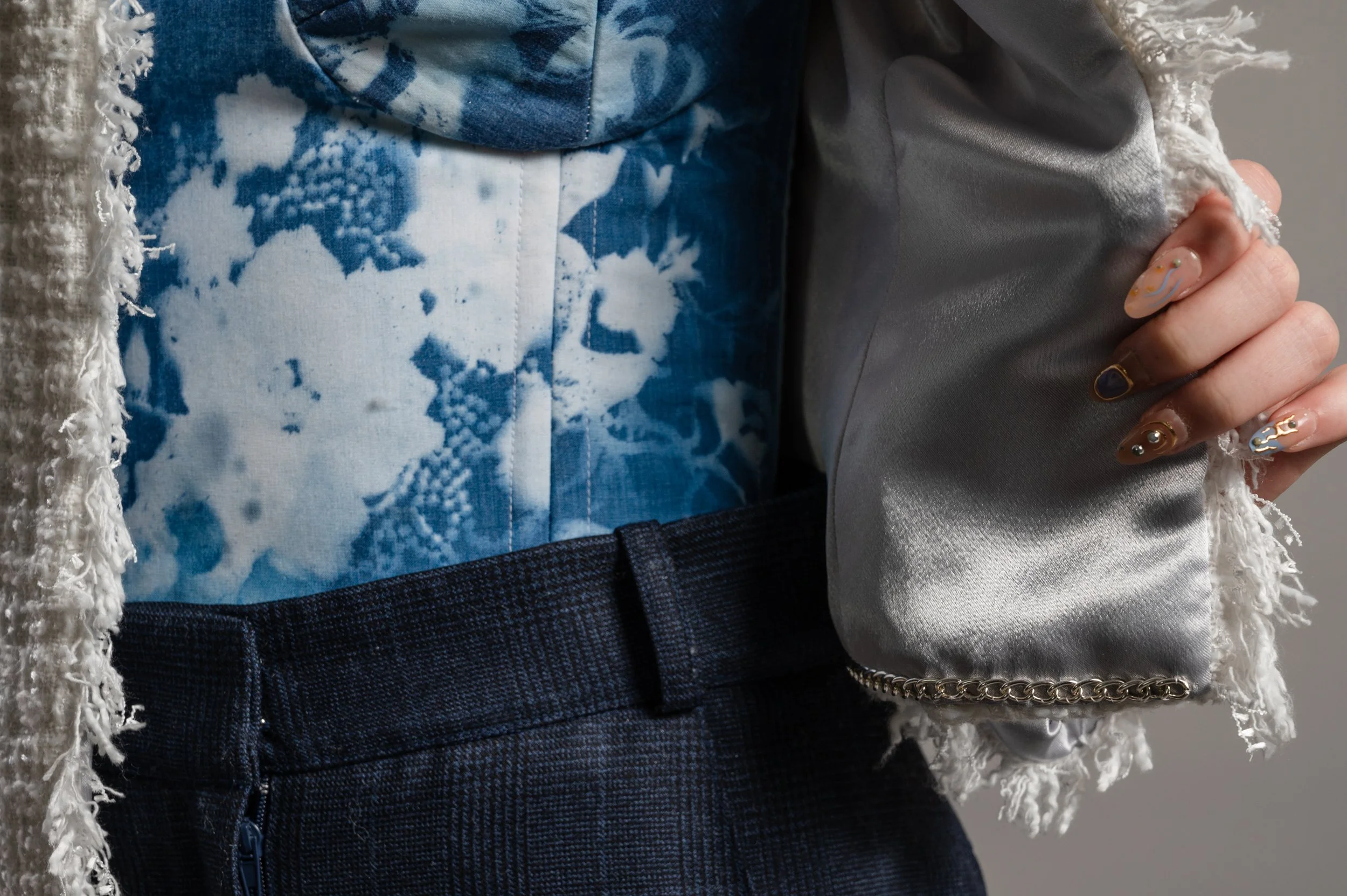 Close-up of a person wearing a white tweed jacket with a grey satin lining, and a corset under dyed with cyanotype. Their hand is holding the jacket.