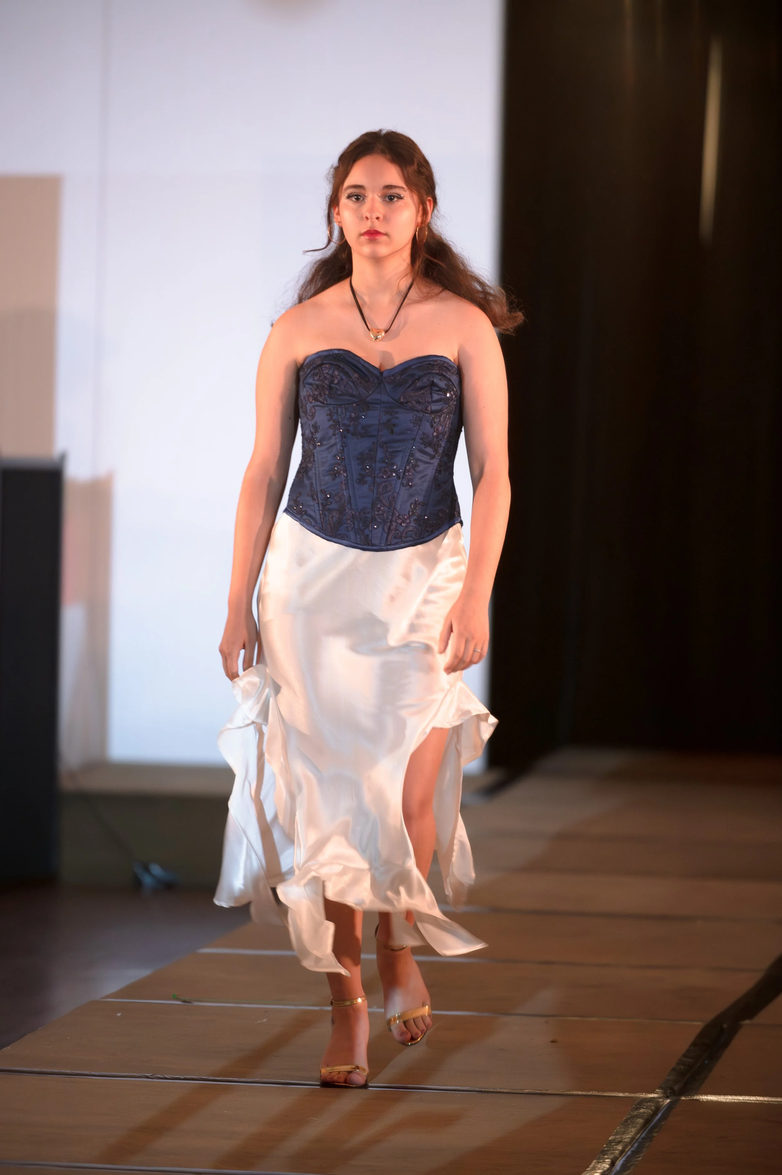 A woman walking on a runway, wearing a strapless navy blue lace corset top and a cream-colored satin skirt, with gold high heels and a necklace.