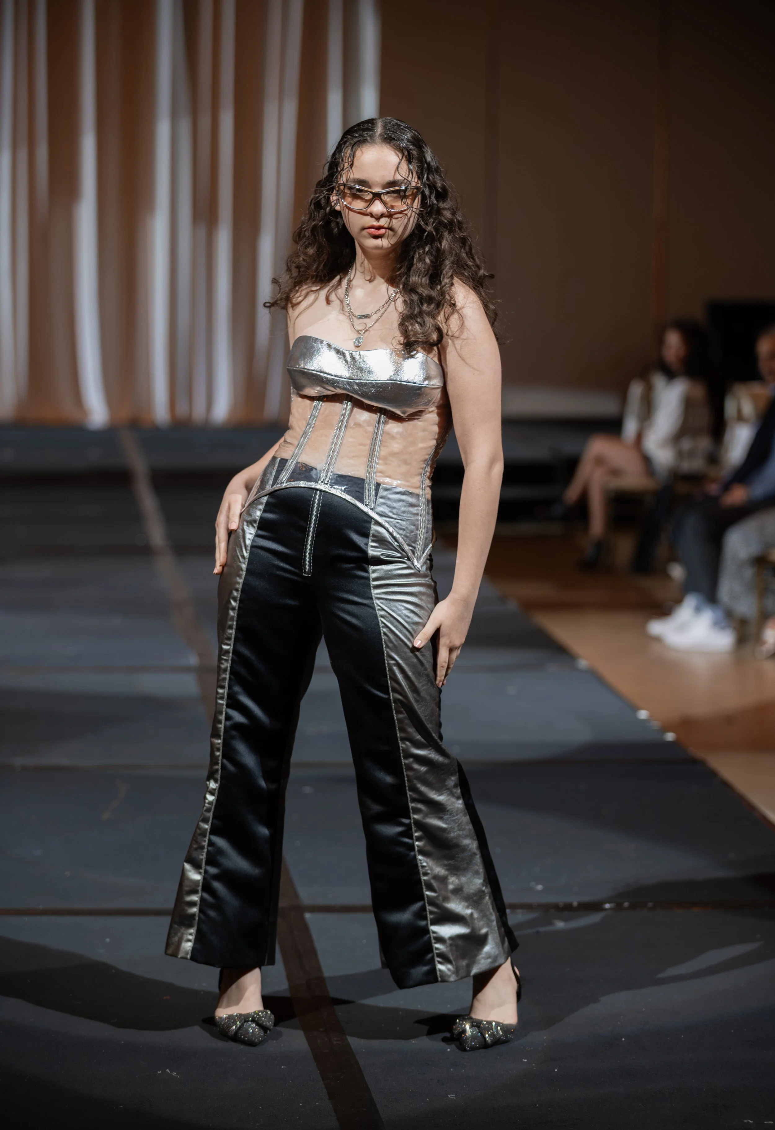 A woman with curly hair and glasses is walking on a runway in a silver and beige strapless top and black wide-leg pants with silver accents. She is wearing slide sandals with embellishments and striped layered necklaces. The background features beige curtains and seated audience members.