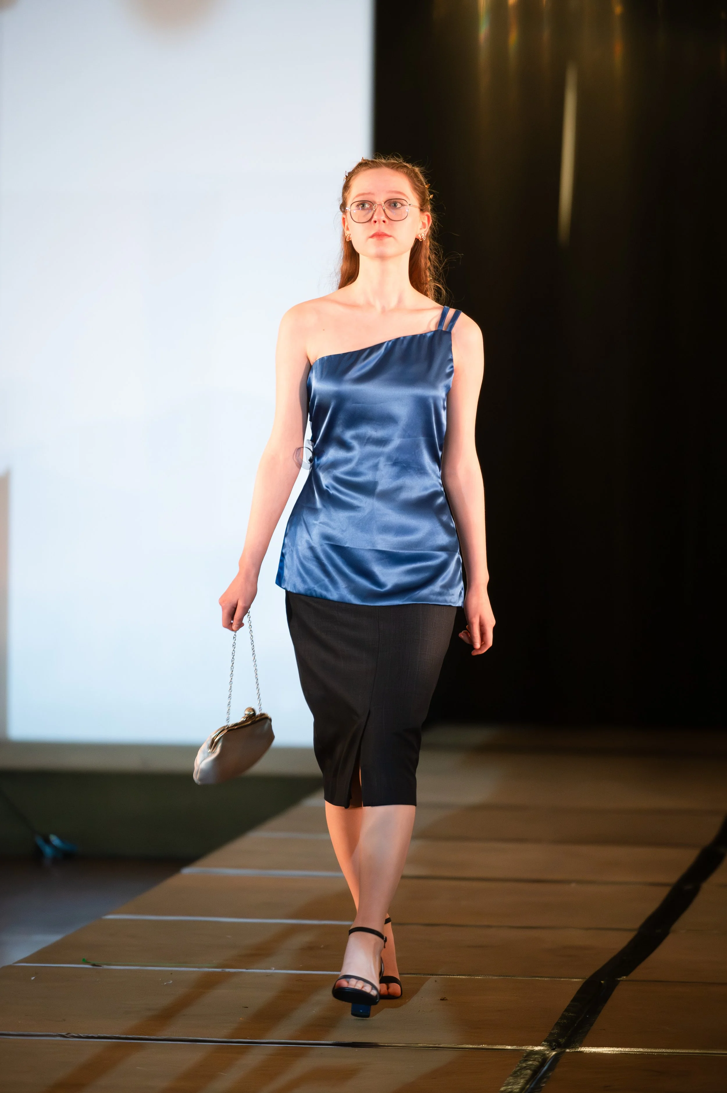 A woman with red hair, wearing glasses, a shiny royal blue satin top with straps, a black skirt, and black heels walks on a runway holding a small bag with a chain strap.