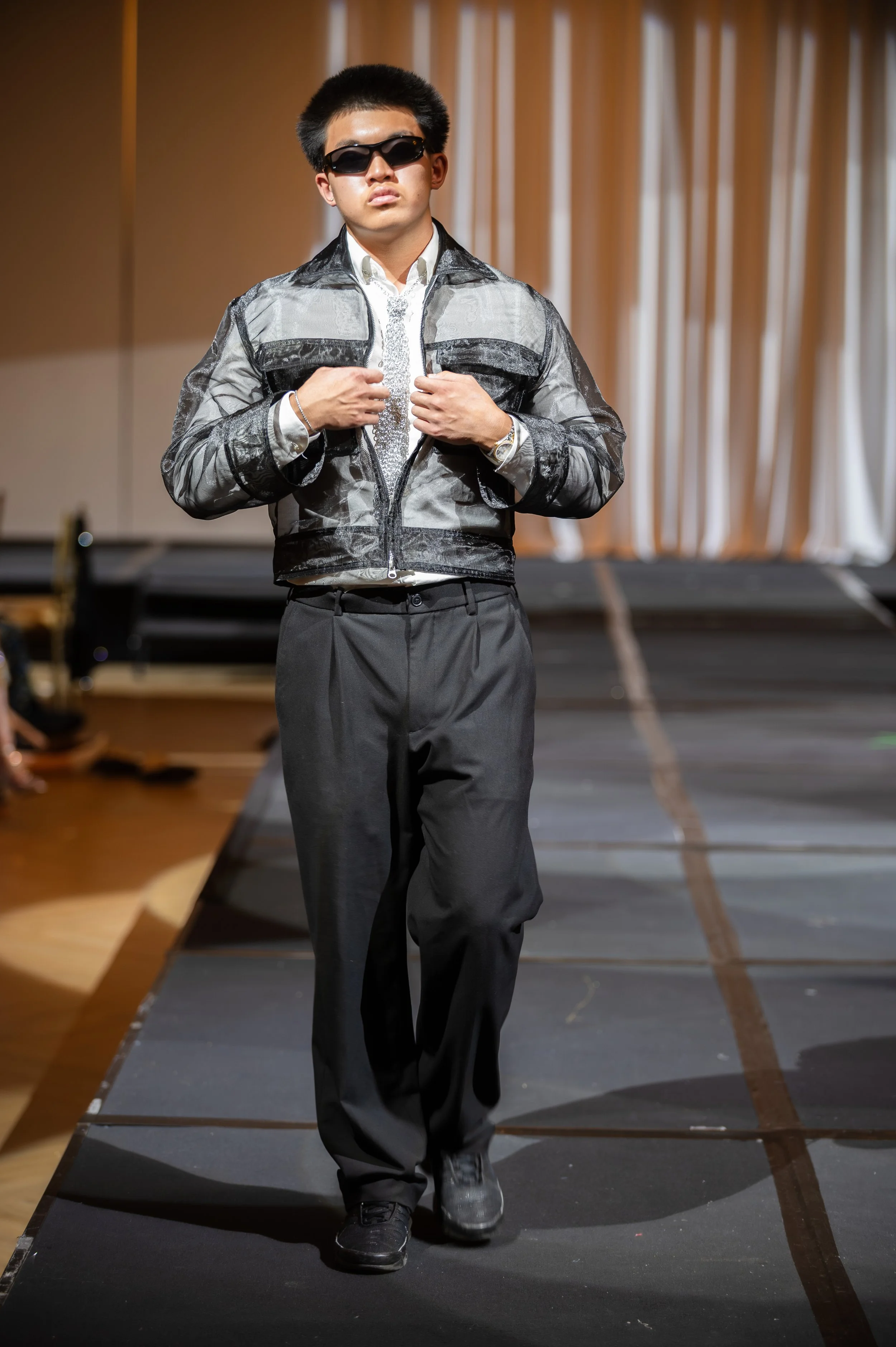 A young man with short black hair wearing dark sunglasses, a sheer black jacket over a white shirt, black pants, and black sneakers walking on a runway during a fashion show with beige curtains in the background.
