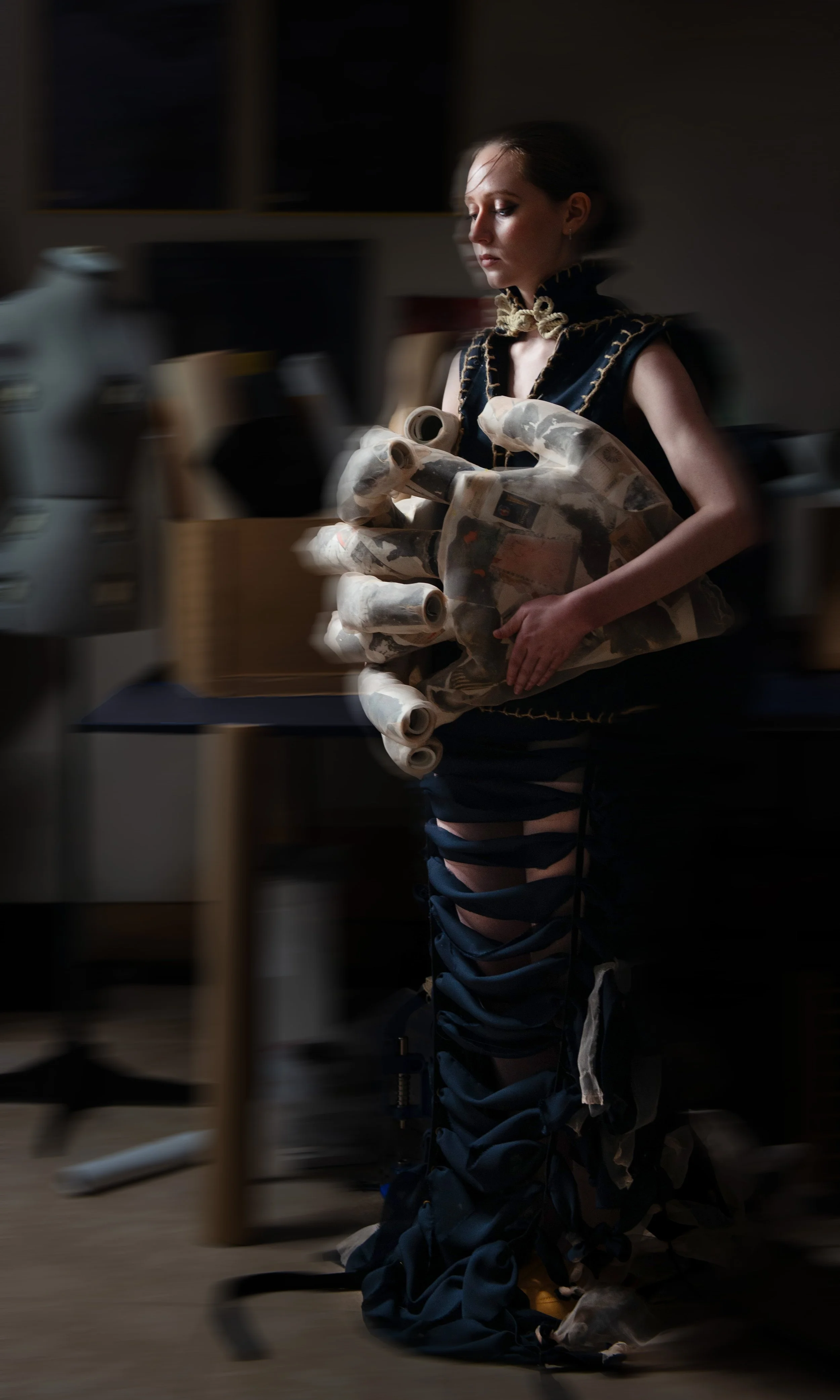 Woman in dark clothing holding a hand-shaped structure, standing in a dimly lit room with workshop or studio equipment.
