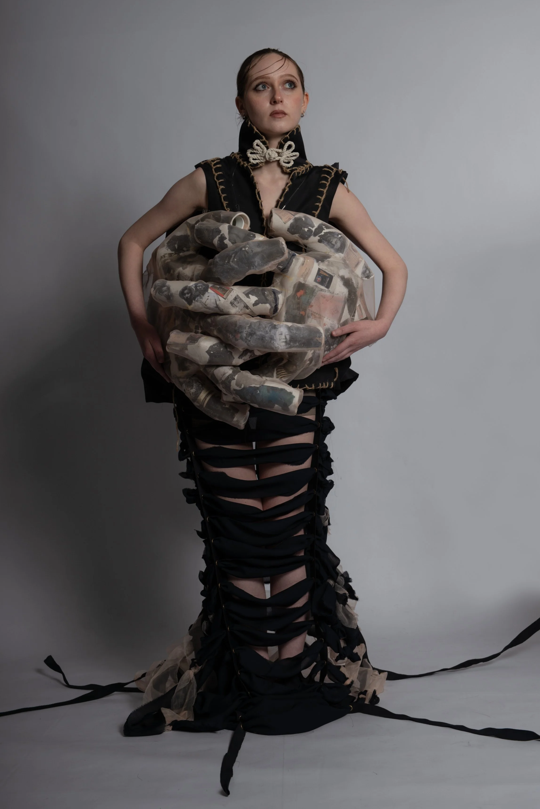 A woman with dark hair styled back, wearing a black outfit with gold-stitch detailing, holds a large, sculptural, hand-shaped object made of collage-like material featuring black and white images, standing against a plain light gray background.