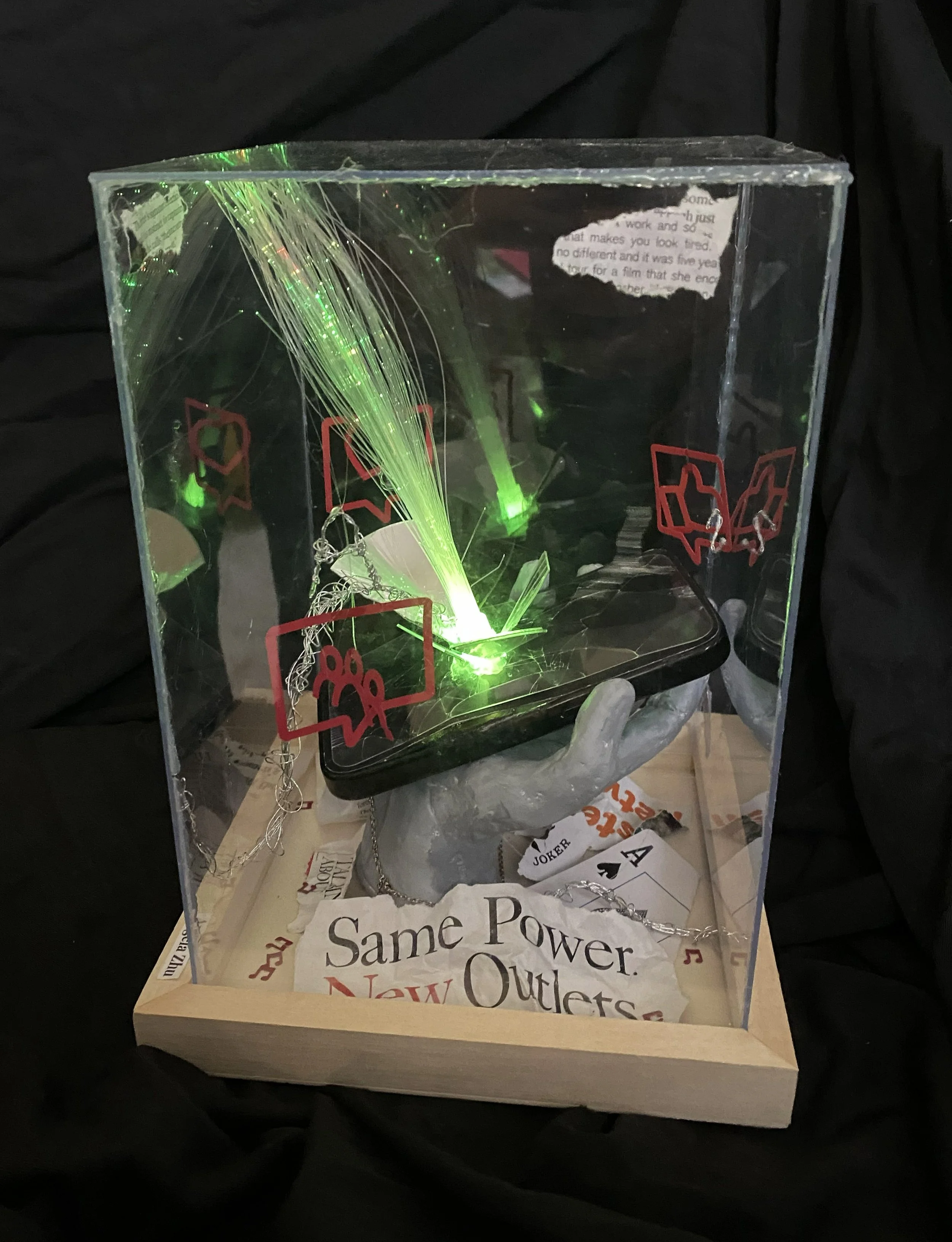 A display case with blue fiber optic branches illuminated, exploding from a phone, held by a clay hand, surrounded by paper with text, on a wooden stand.