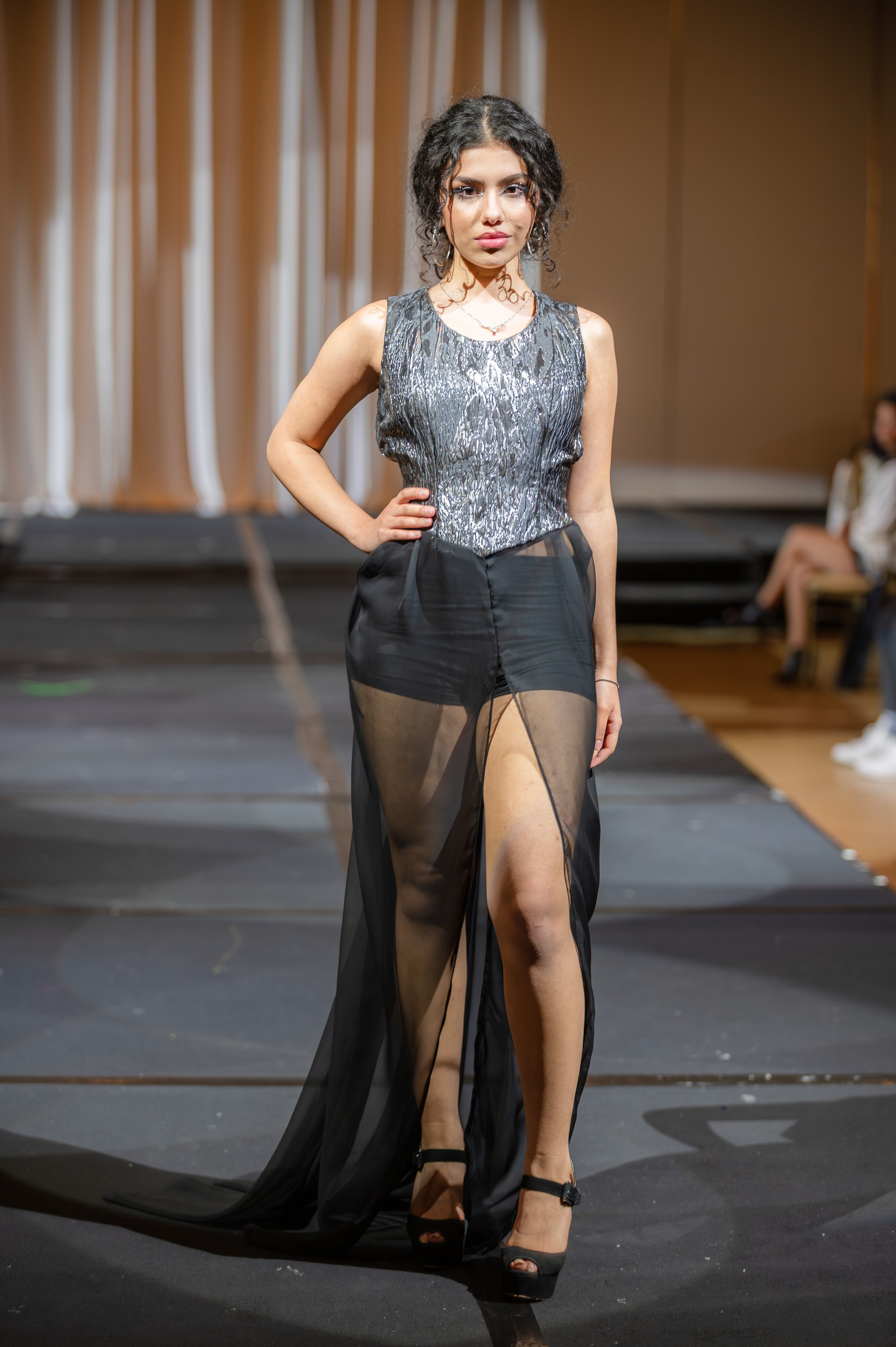 A female model walking on a runway, wearing a sleeveless silver top with a shiny pattern and a sheer black skirt with high slits, in an indoor fashion show setting.