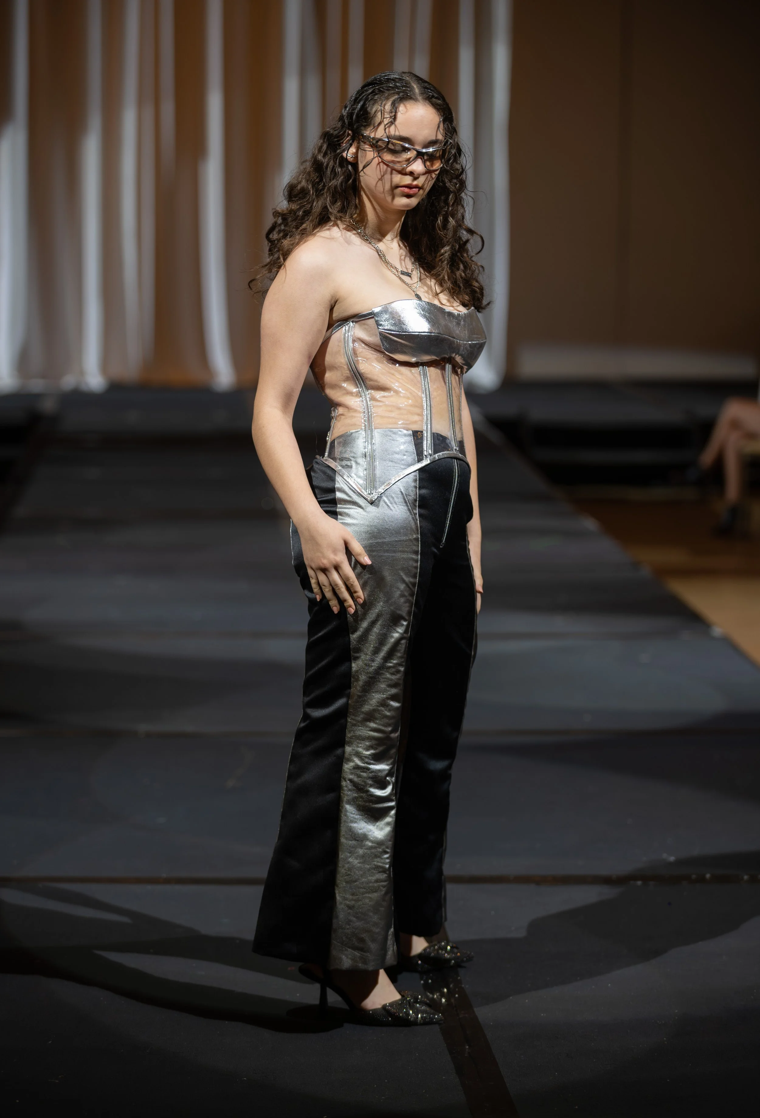 A woman with curly hair and wearing glasses stands on a stage, dressed in a metallic and sheer strapless top with matching shiny black pants and high heels.