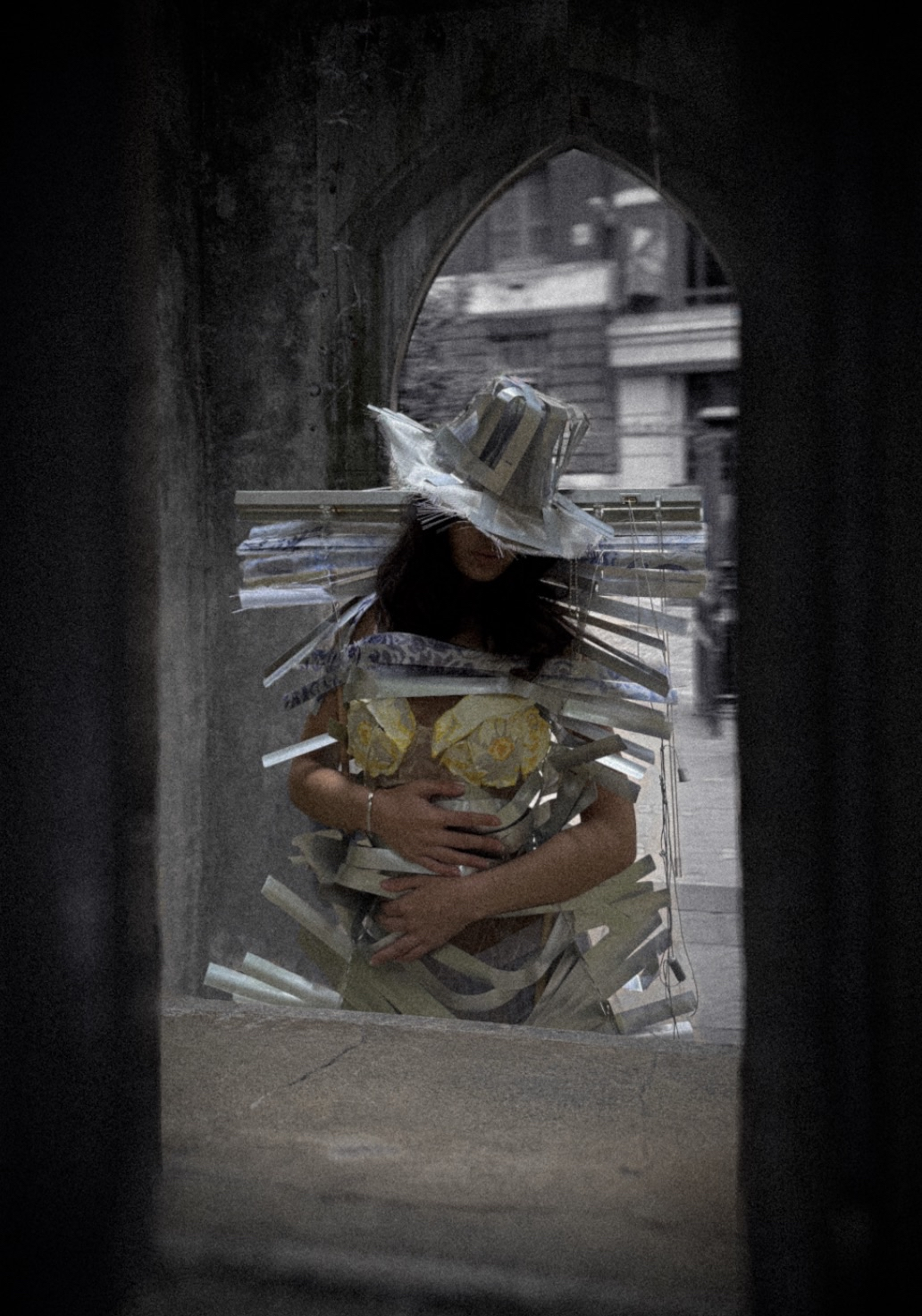 Model wearing garment and hat made out of aluminium window blinds, with yellow floral bra top peaking from under. Standing through a small opening between stone structures as part of an artistic or fashion photoshoot.