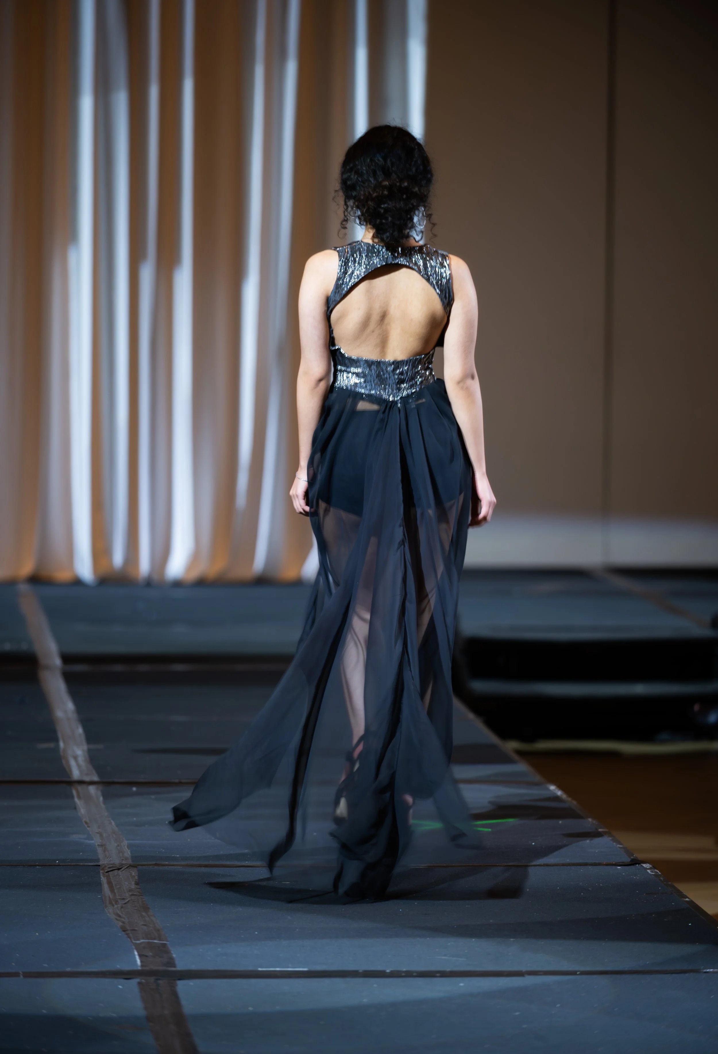Back view of a woman in an elegant black dress with a large open back, walking on a runway with a curtain backdrop.
