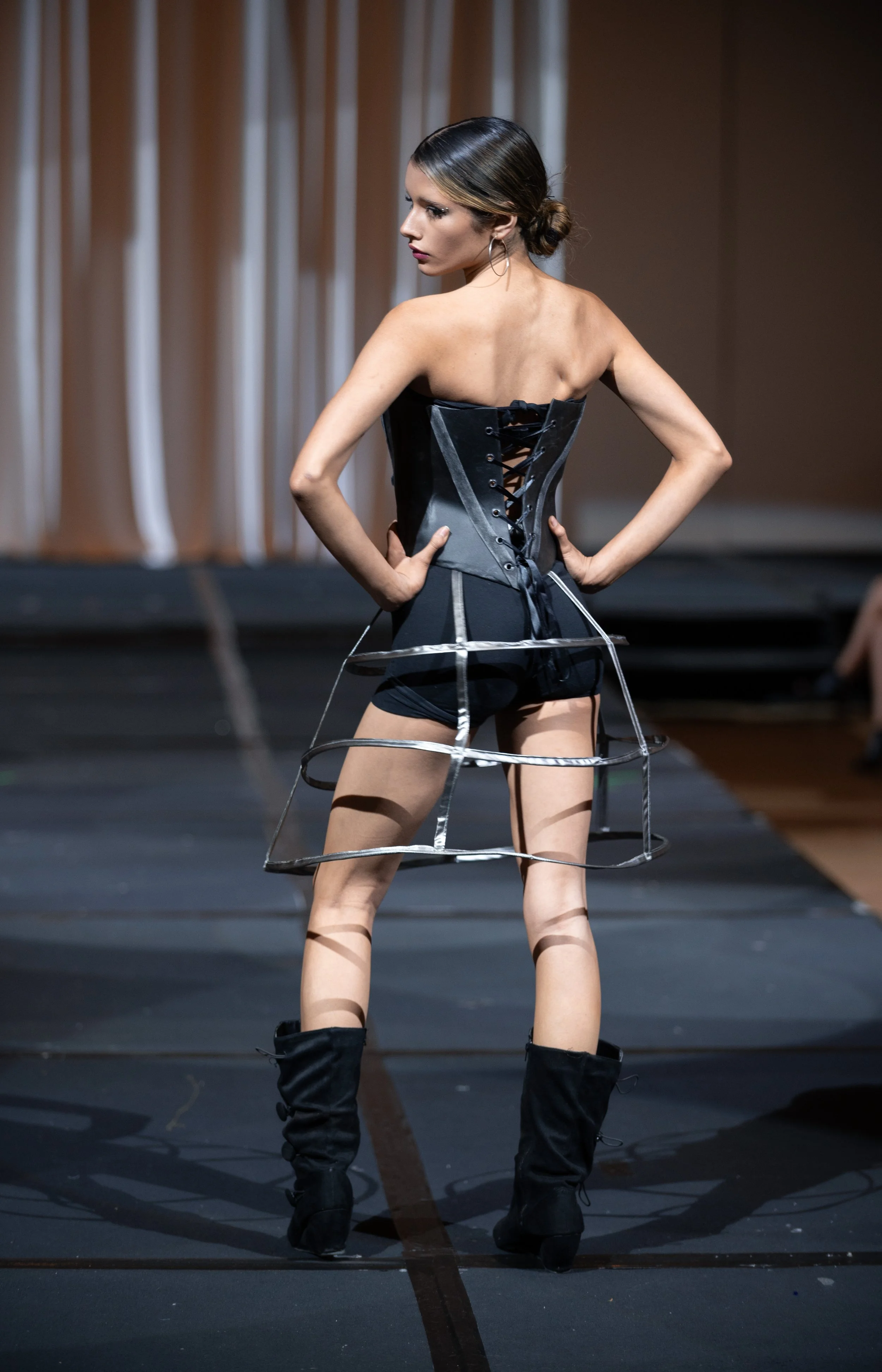 A woman on a runway wears a black corset-style top with lace-up back and a skirt with circular metal frame. She has dark hair styled in a low bun and is wearing hoop earrings and black knee-high boots.