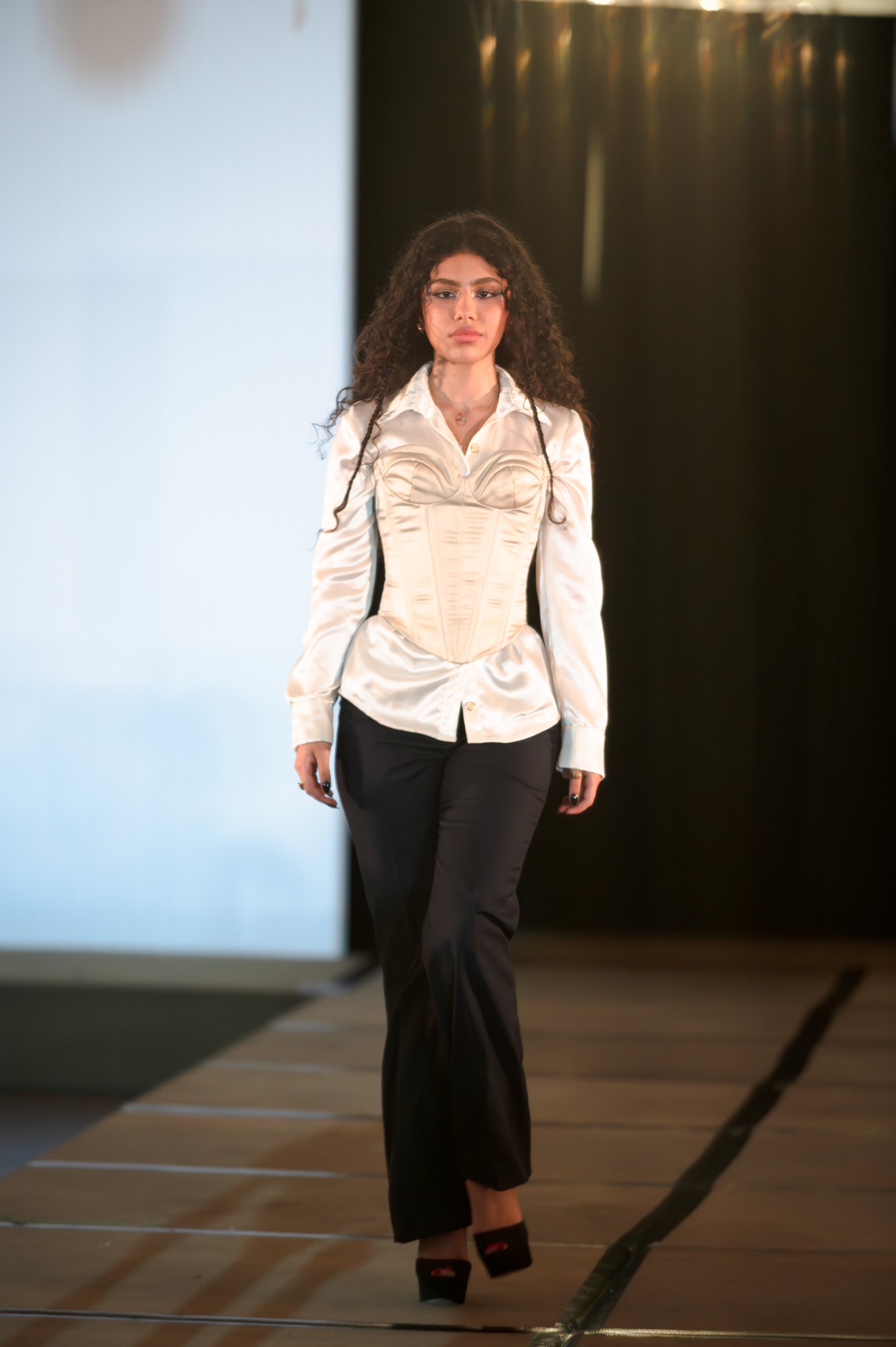 A woman with curly hair walking on a runway during a fashion show, wearing a cream-colored corset-style top and black pants, with black high-heeled shoes.