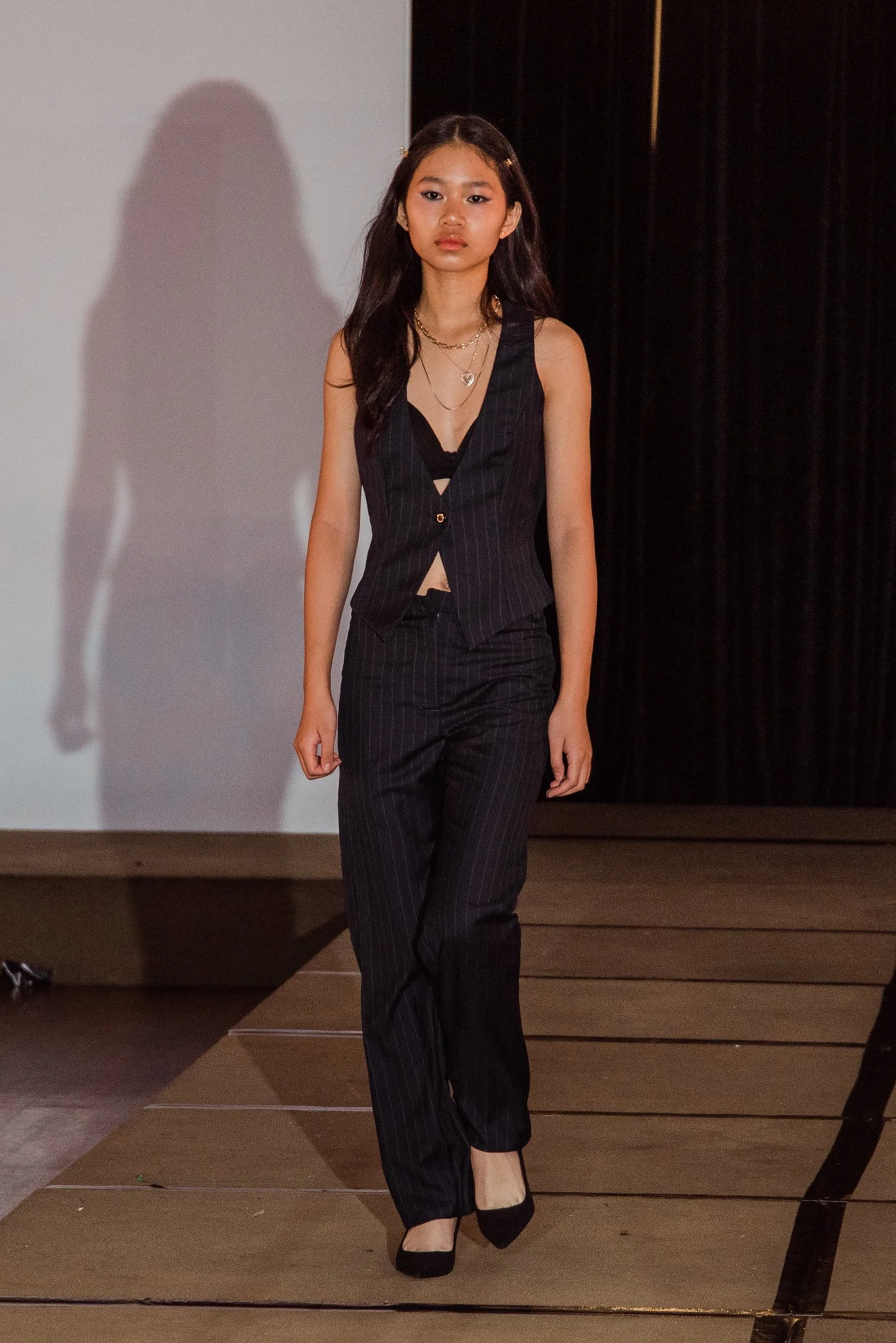 A young woman walking on stage in black pinstripe suit, black heels, and gold jewelry, with a neutral expression.