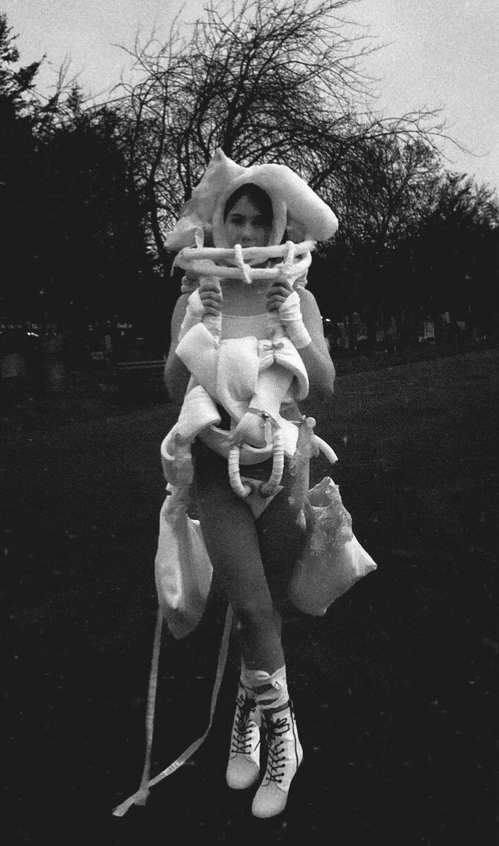 A person wearing white avant-garde clothing, including high-heeled boots, gloves,, standing outside at dusk with trees and park benches in the background.